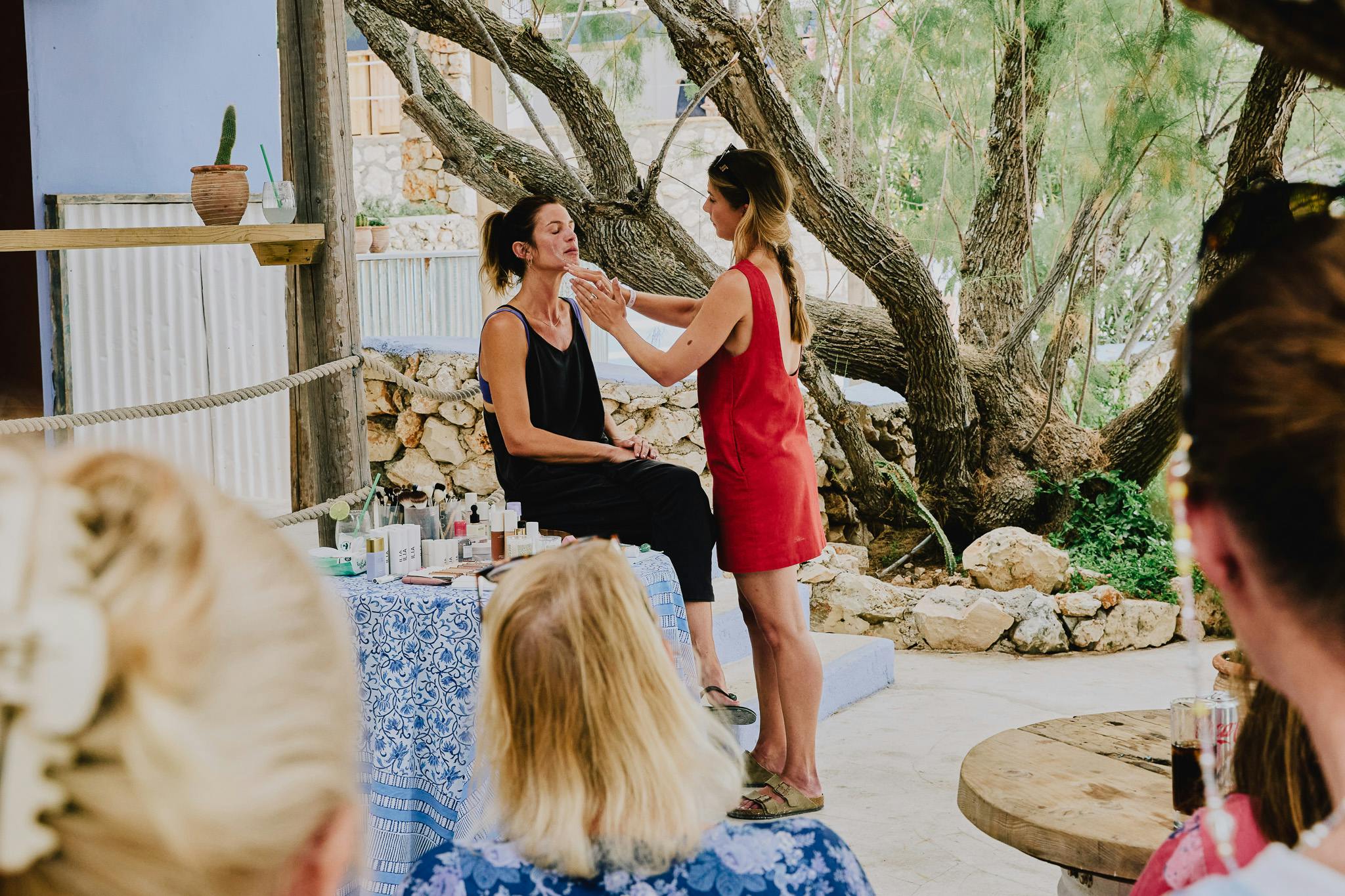 A woman in a red dress demonstrates beauty treatments on a woman in black, set in the rustic surrounds of a Mediterranean island. There is a table covered in products and a blue patterned tablecloth. You can see the back of guests heads who are watching the demonstration.