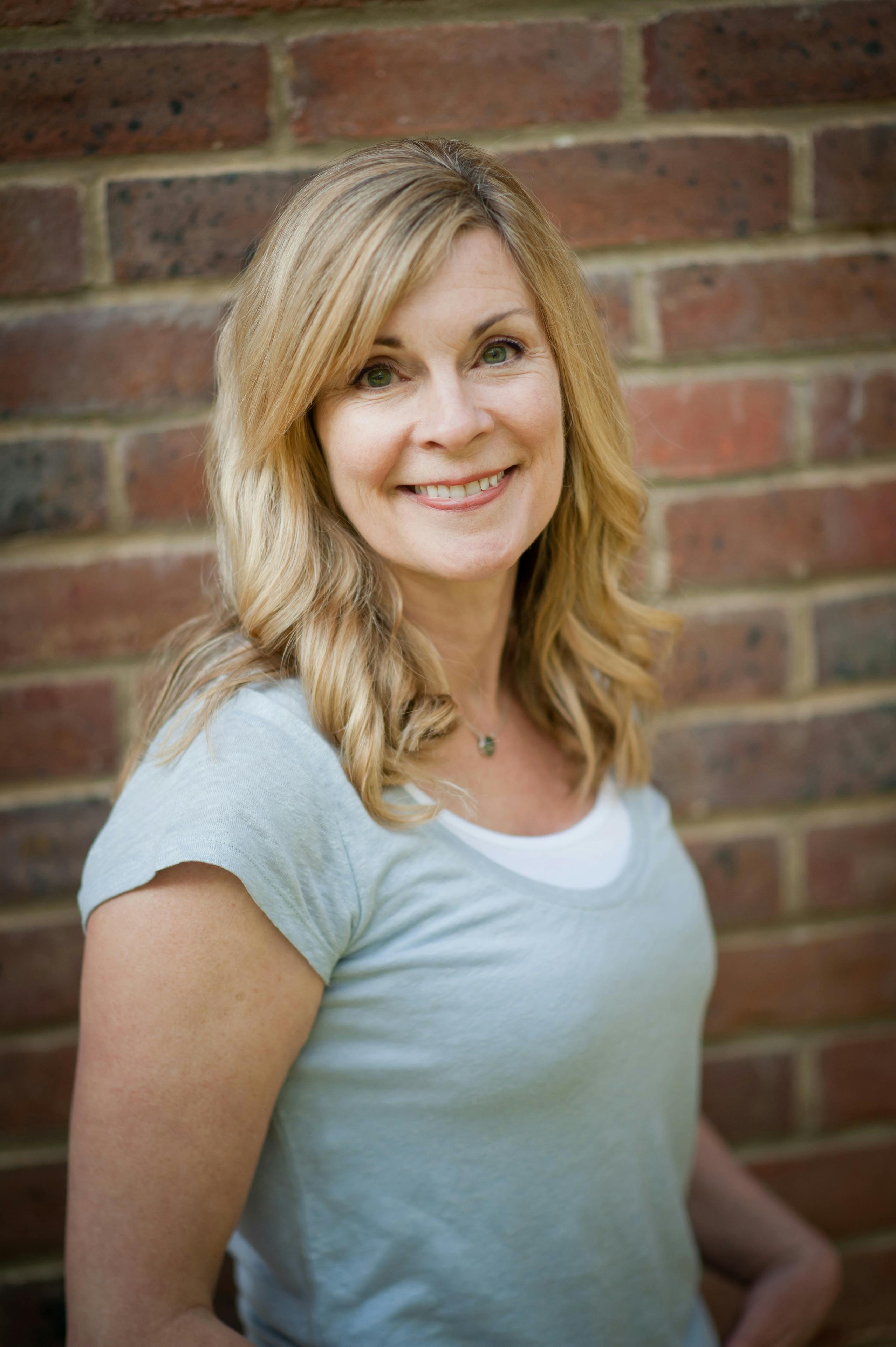 A woman with blonde hair, wearing a grey T-shirt, stands in front of a brick wall, smiling into the camera.
