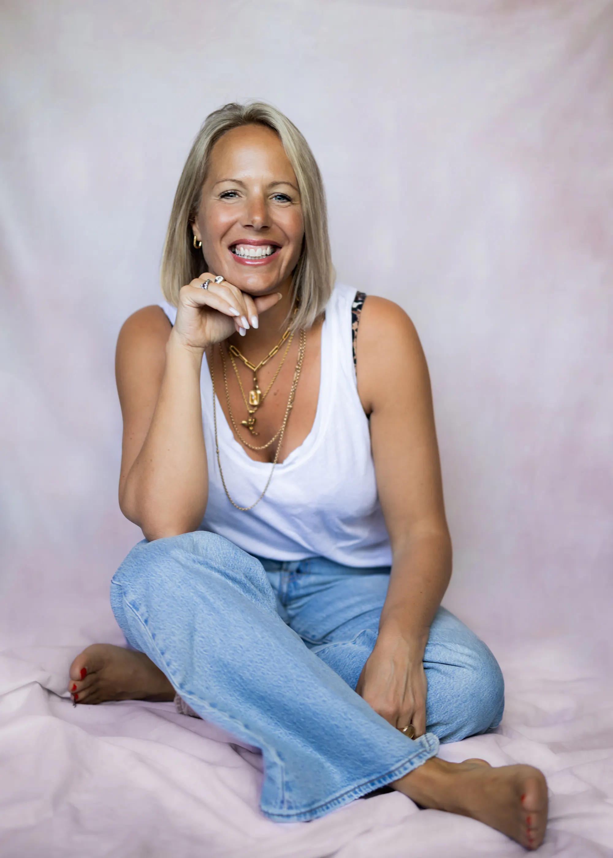 Woman with blonde hair in white tank top and blue jeans and barefoot, on a white professional photography backdrop, looking at the camera and smiling, resting her chin against her hand.