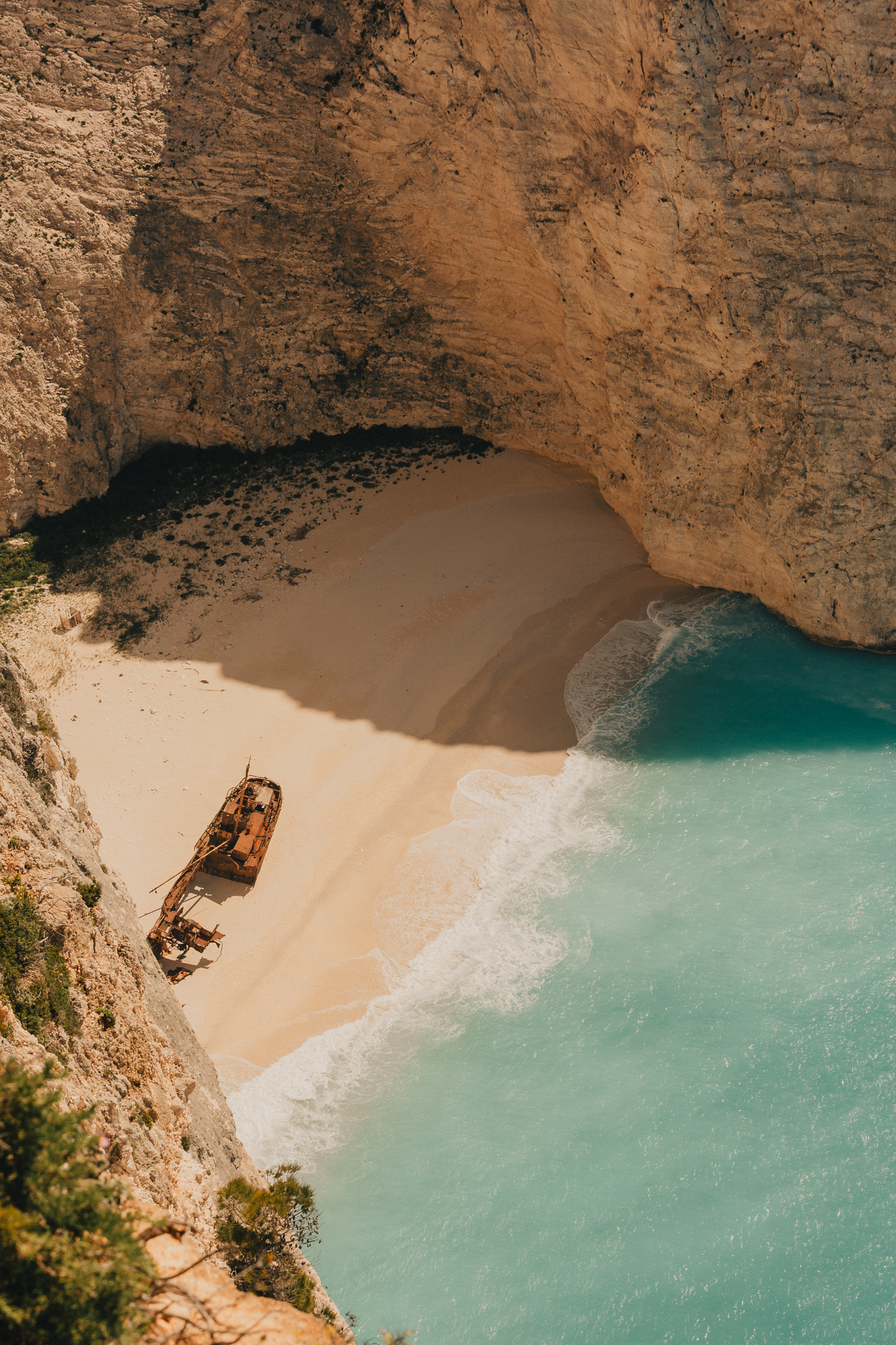 A rusted shipwreck lies stranded on a secluded sandy cove, encircled by towering golden limestone cliffs and lapped by vivid turquoise water. Shot from high above, the image has an otherworldly, mythic quality — beautiful and eerie in equal measure.