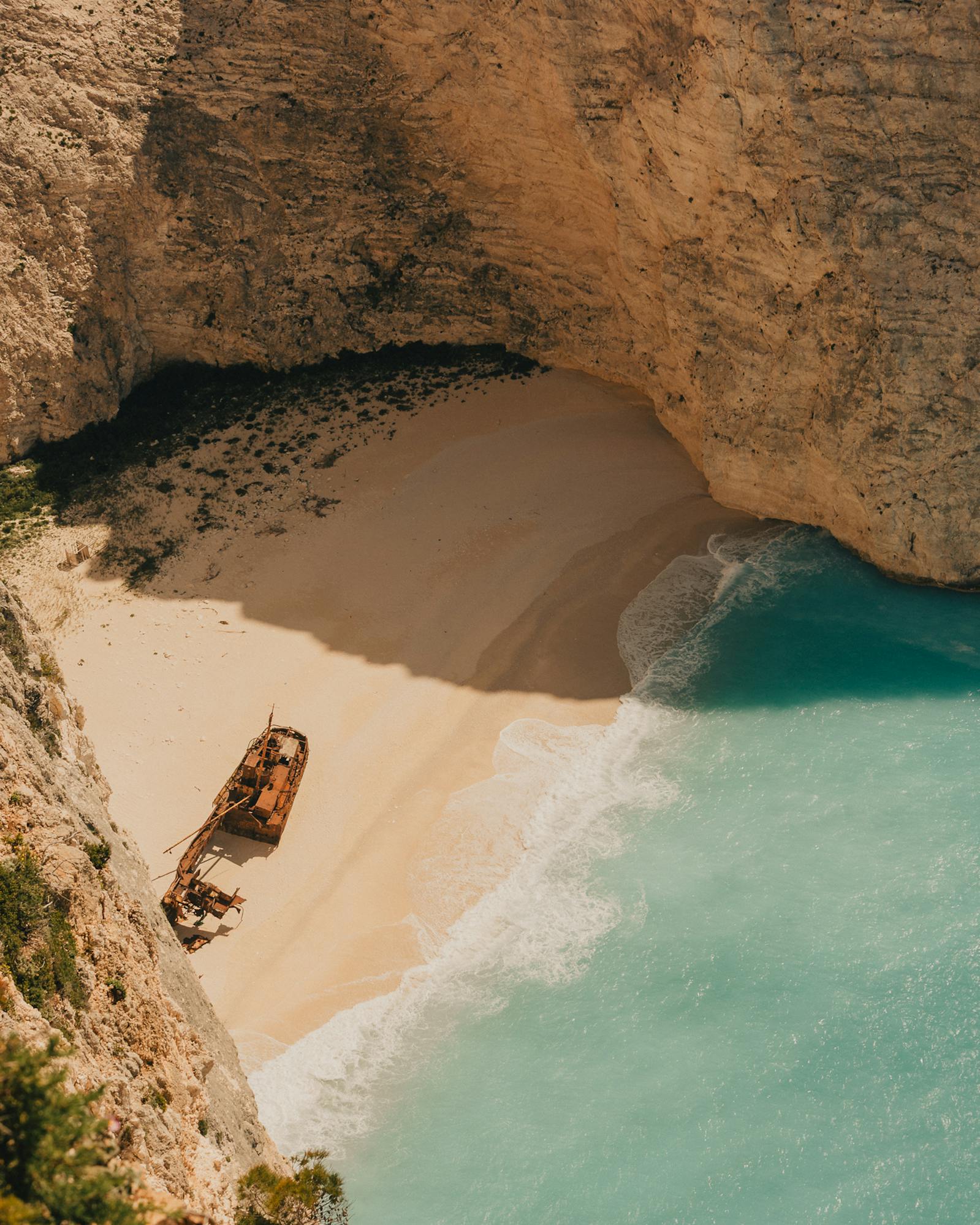 A rusted shipwreck lies stranded on a secluded sandy cove, encircled by towering golden limestone cliffs and lapped by vivid turquoise water. Shot from high above, the image has an otherworldly, mythic quality — beautiful and eerie in equal measure.