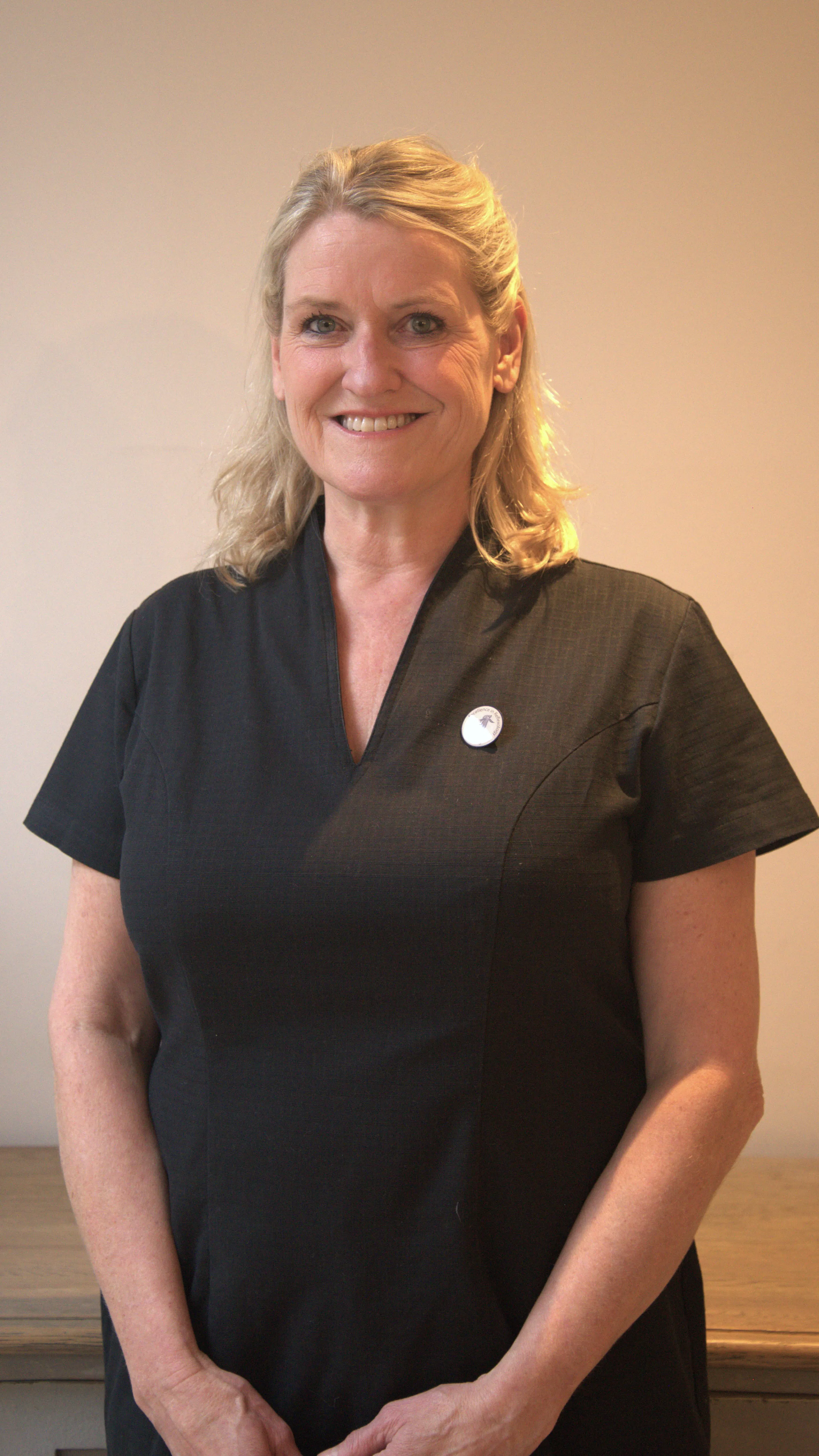 A blonde woman is standing with her hands linked looking at the camera and smiling wearing a black top.