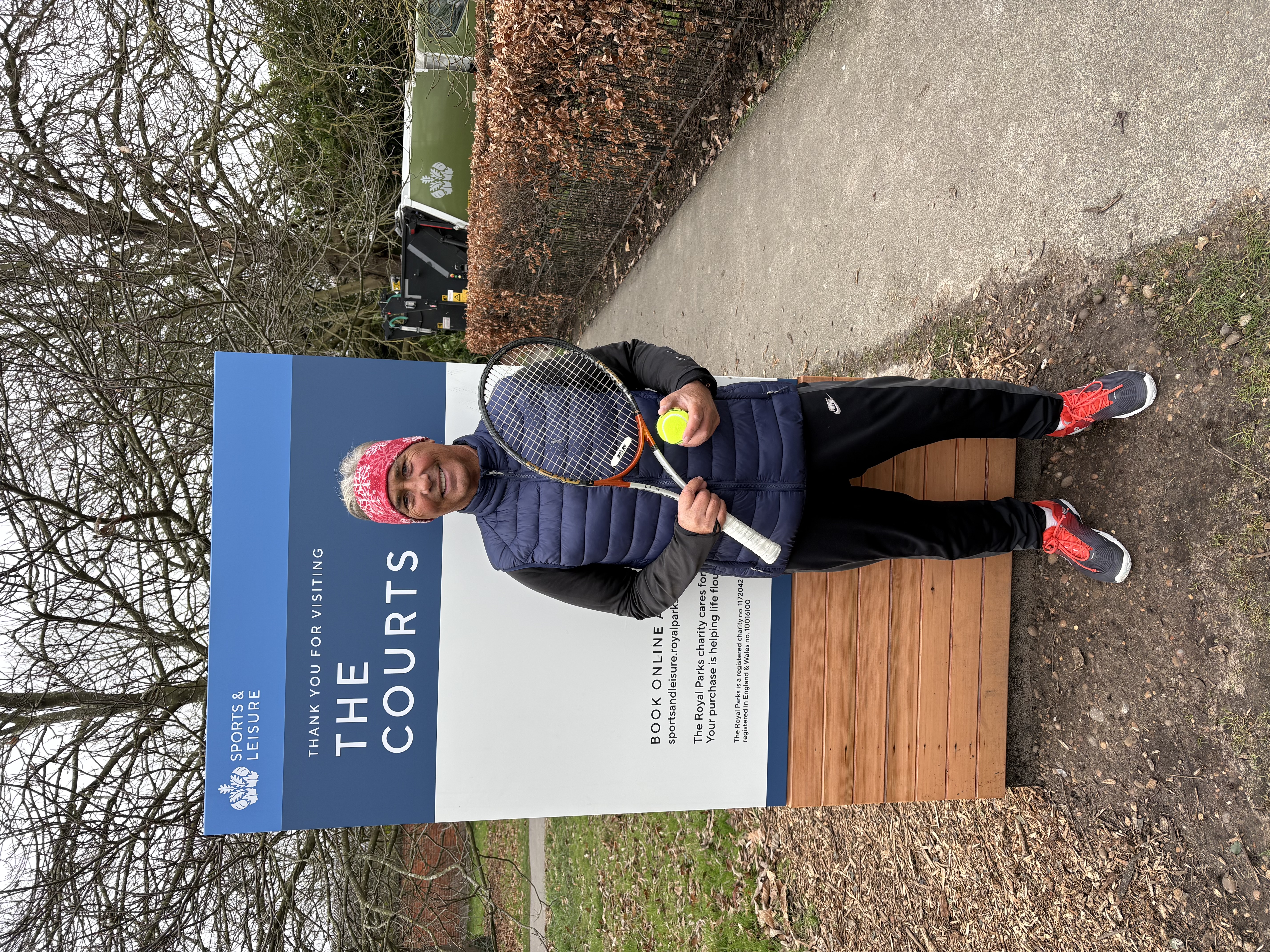 A man standing in a park in front of a 'the courts' billboard and holding a tennis racket with a tennis ball in sportswear and a red bandana. 