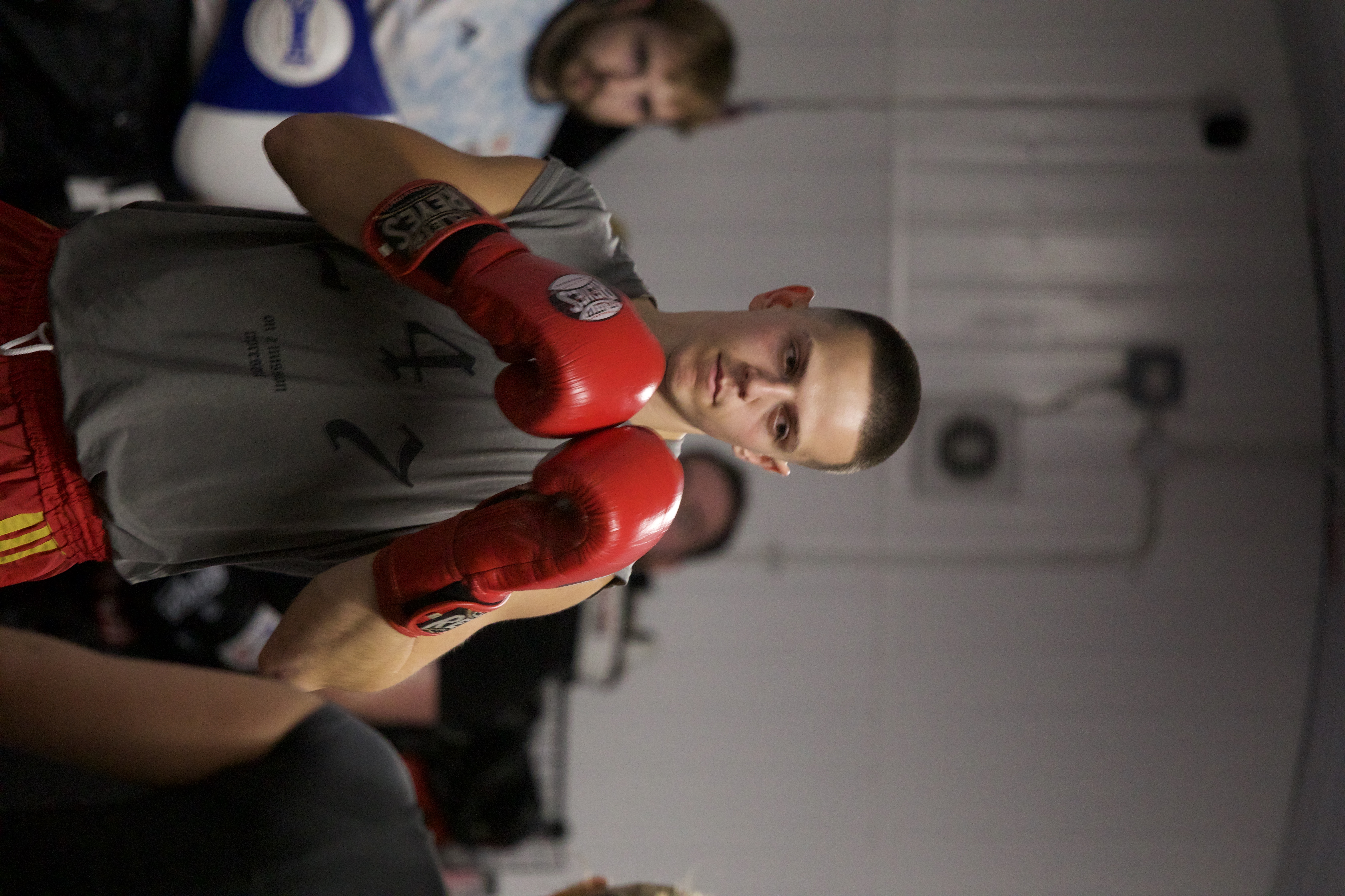 A man in red boxing gloves and a grey tank top with a serious look on his face, getting ready for a spar.