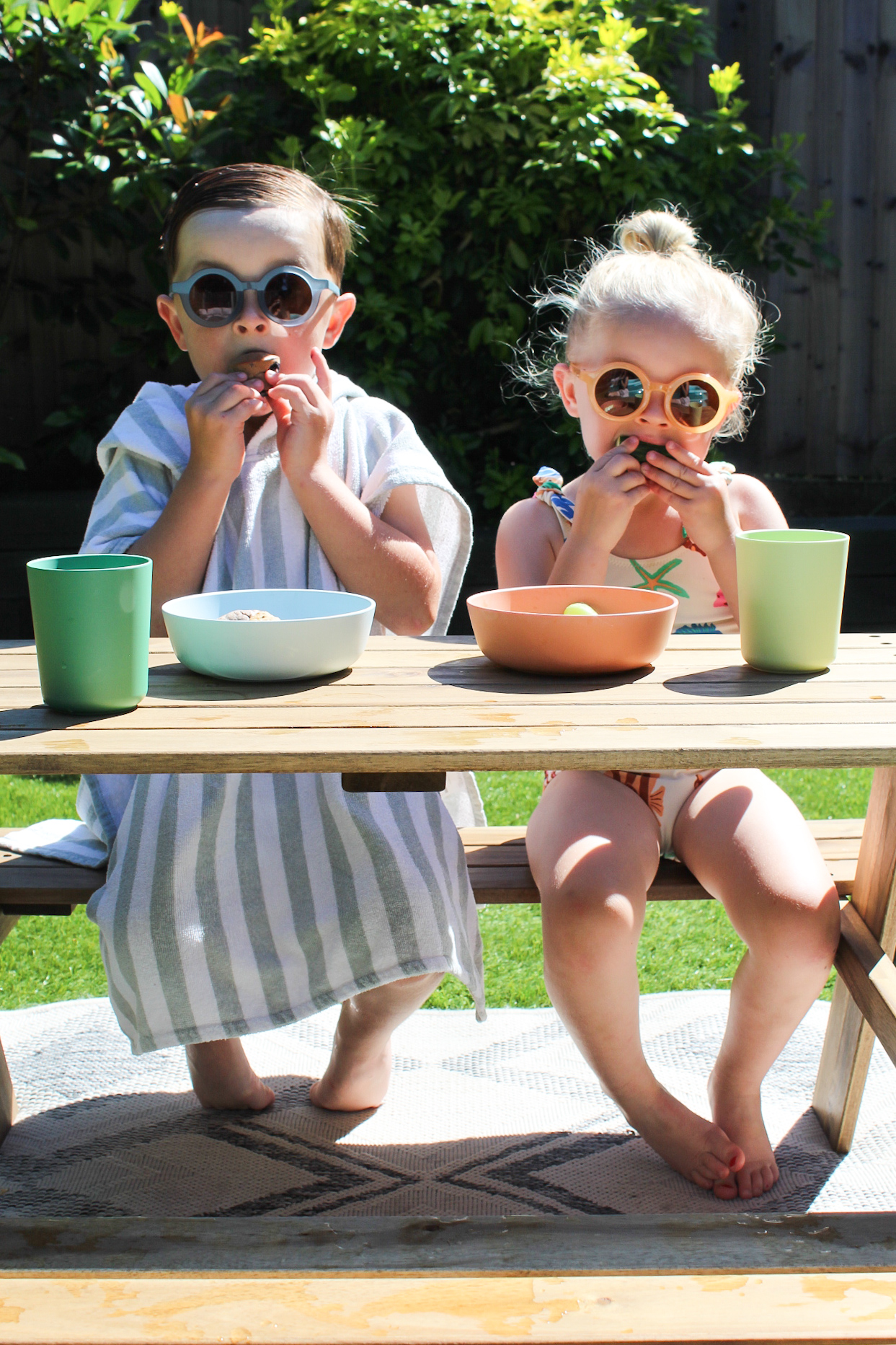 Two small toddlers sit side by side at a low wooden picnic table in a bright sunny garden. Both are wearing round, retro-style sunglasses — one in blue-grey, one in yellow — and are mid-bite, snacking with both hands. The table is set with pastel-coloured bowls and cups in mint green and peach. One child is wrapped in a blue and white striped towel, while the other wears a floral swimsuit. The scene is cheerful and candid, capturing a fun, carefree summer moment.