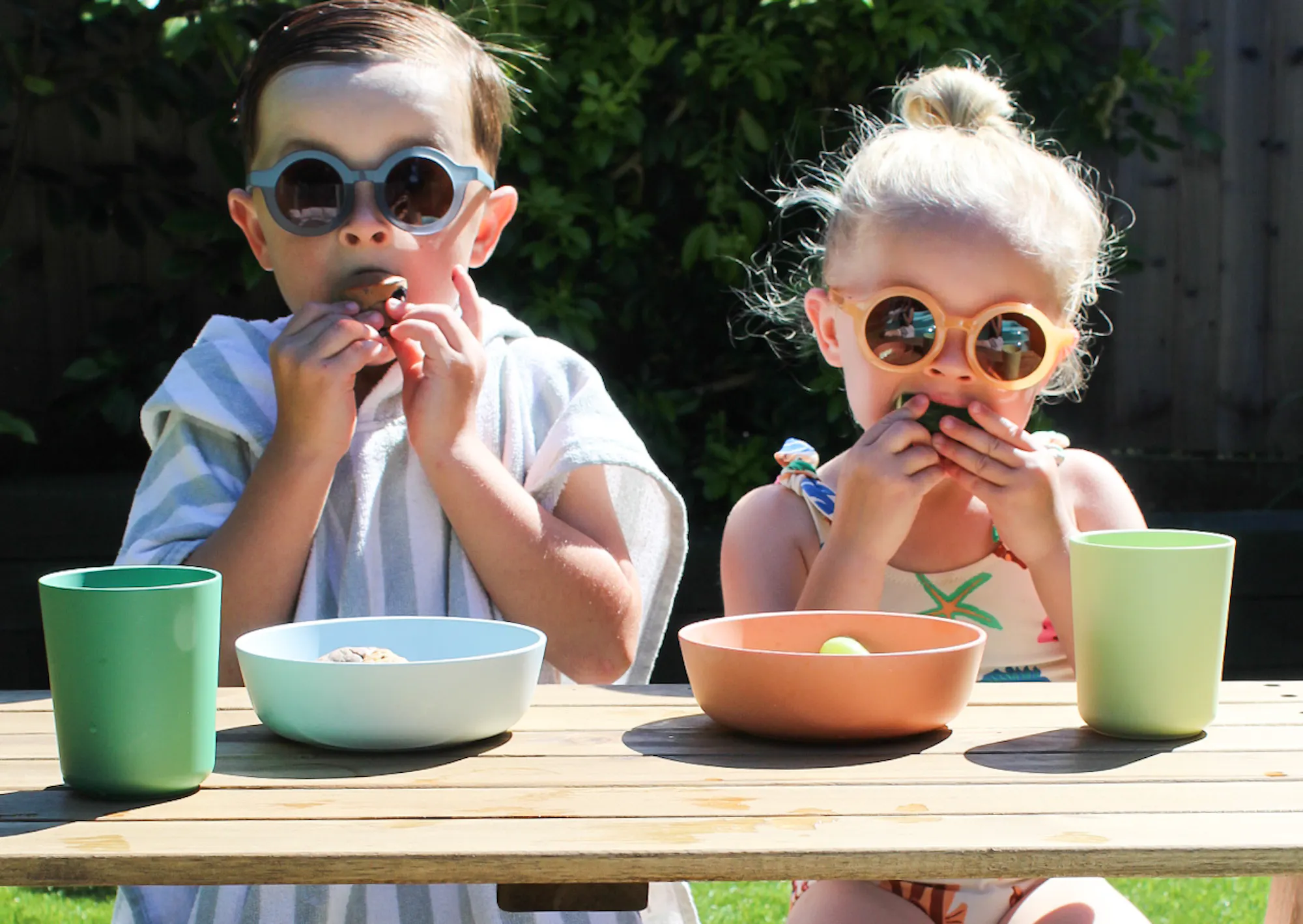 Two small toddlers sit side by side at a low wooden picnic table in a bright sunny garden. Both are wearing round, retro-style sunglasses — one in blue-grey, one in yellow — and are mid-bite, snacking with both hands. The table is set with pastel-coloured bowls and cups in mint green and peach. One child is wrapped in a blue and white striped towel, while the other wears a floral swimsuit. The scene is cheerful and candid, capturing a fun, carefree summer moment.