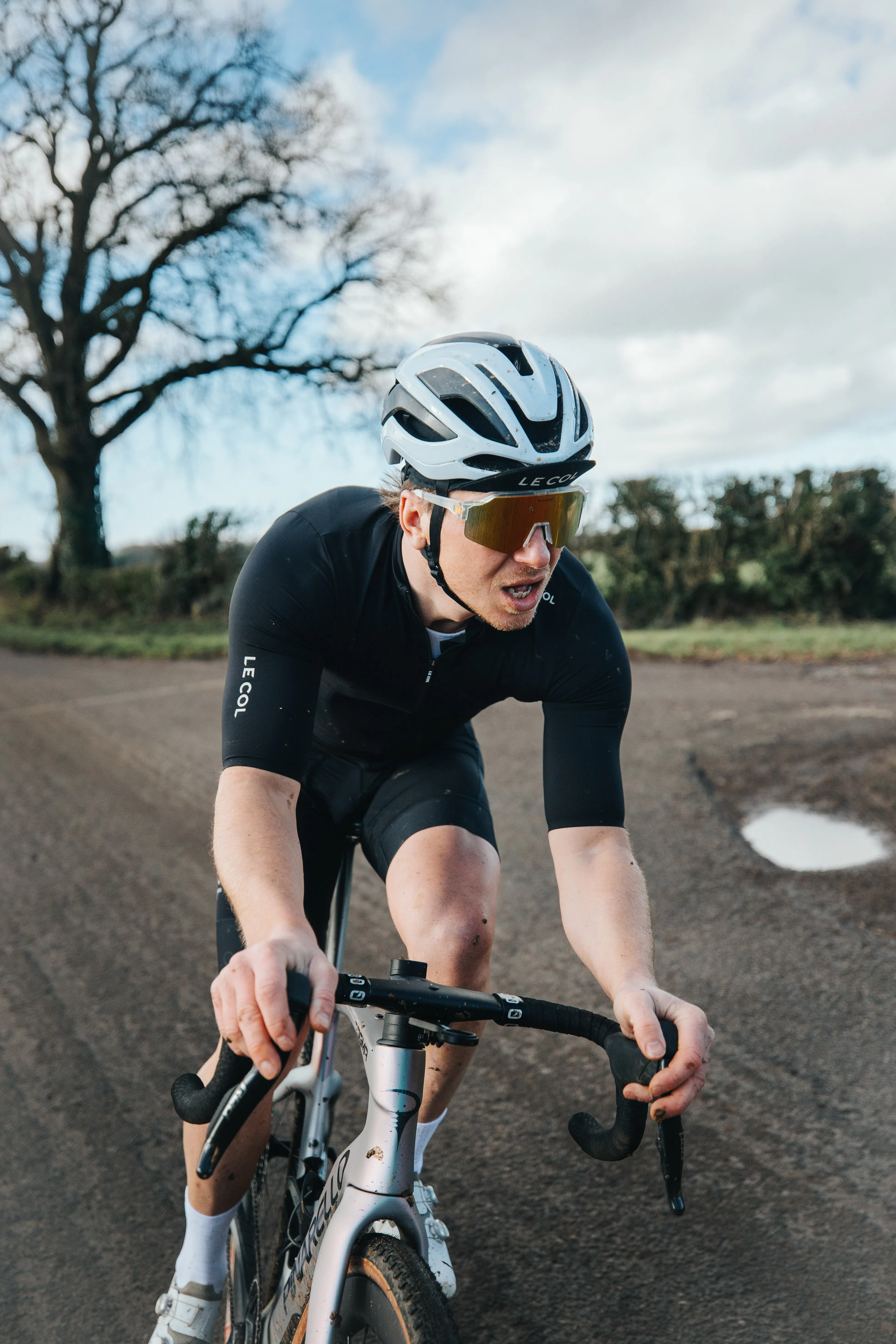 Man in action on a bicycle on a country lane wearing all back and a white helmet.
