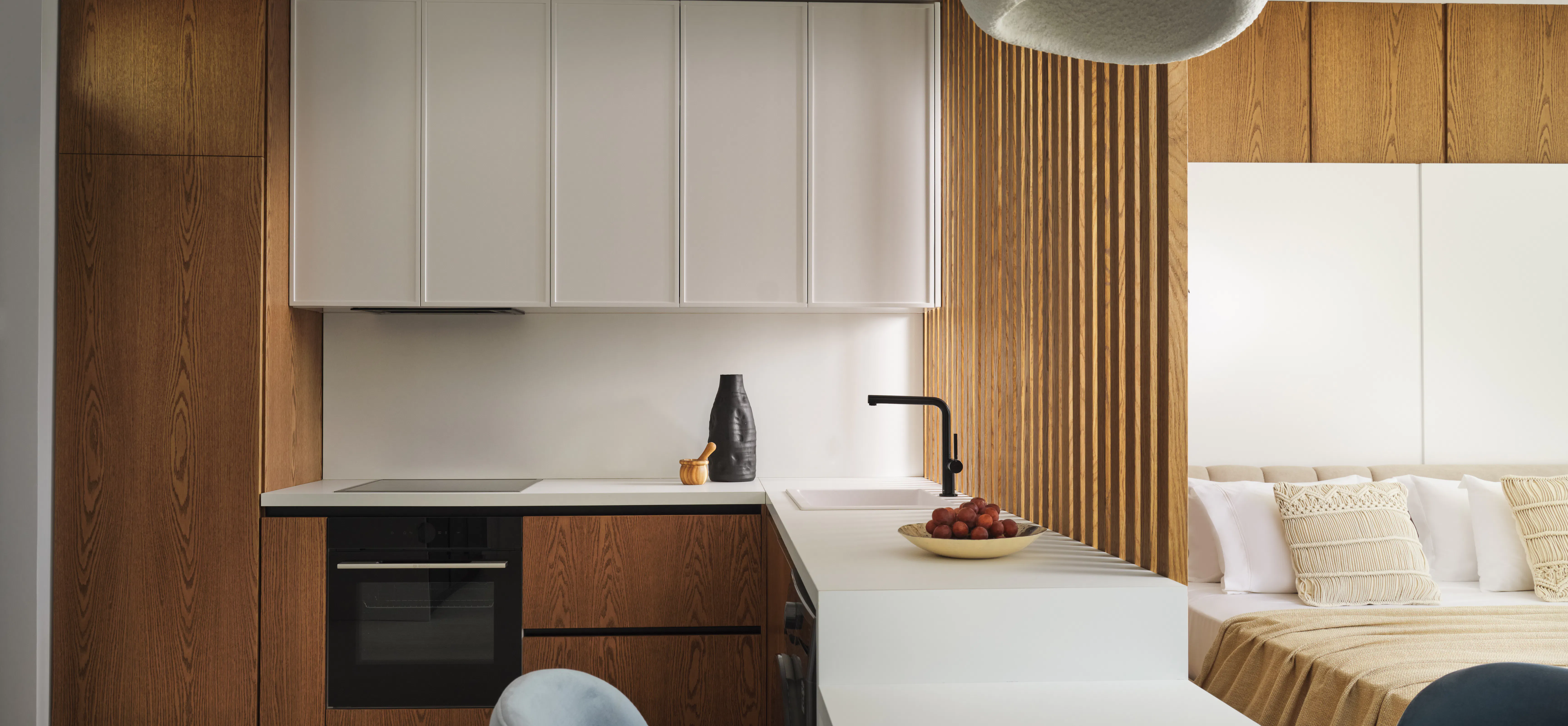 Open-plan kitchenette and bedroom with wood and white cabinetry, white island counter, ribbed wood accent wall, and sculptural pendant light