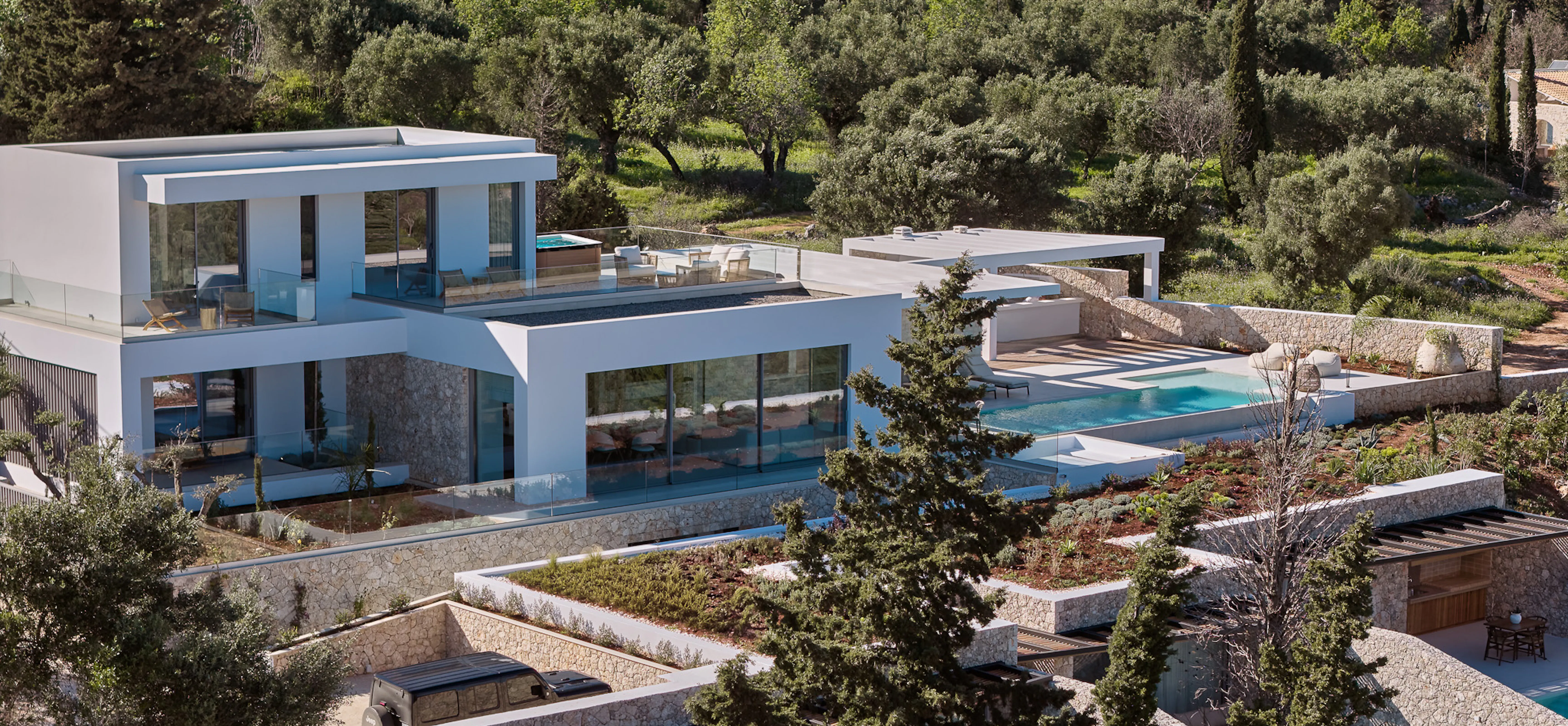 Aerial view of a contemporary white villa cascading down a hillside through dense olive groves, with multiple terraces, pools, and stone retaining walls blending modern architecture into the natural landscape.