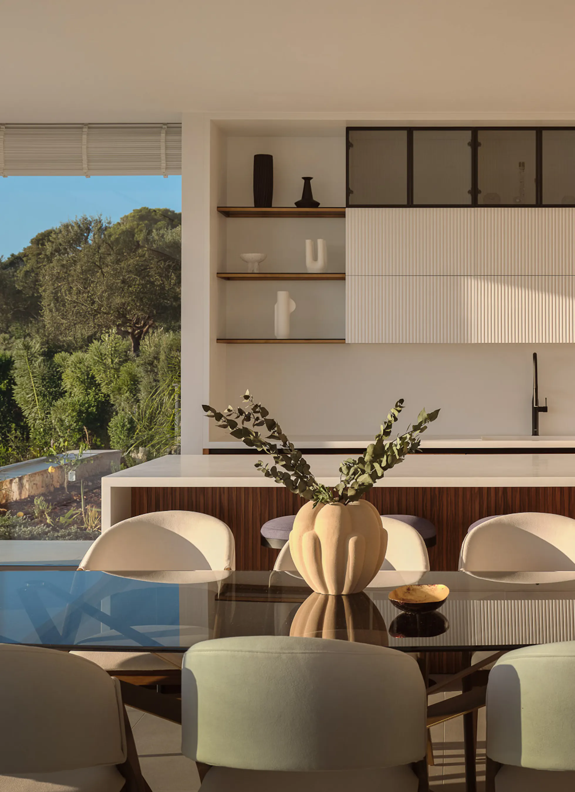 Sophisticated kitchen-dining room with sage green upholstered chairs around a dark oval table, warm walnut cabinetry, fluted white panels, and curated open shelving — refined yet inviting.