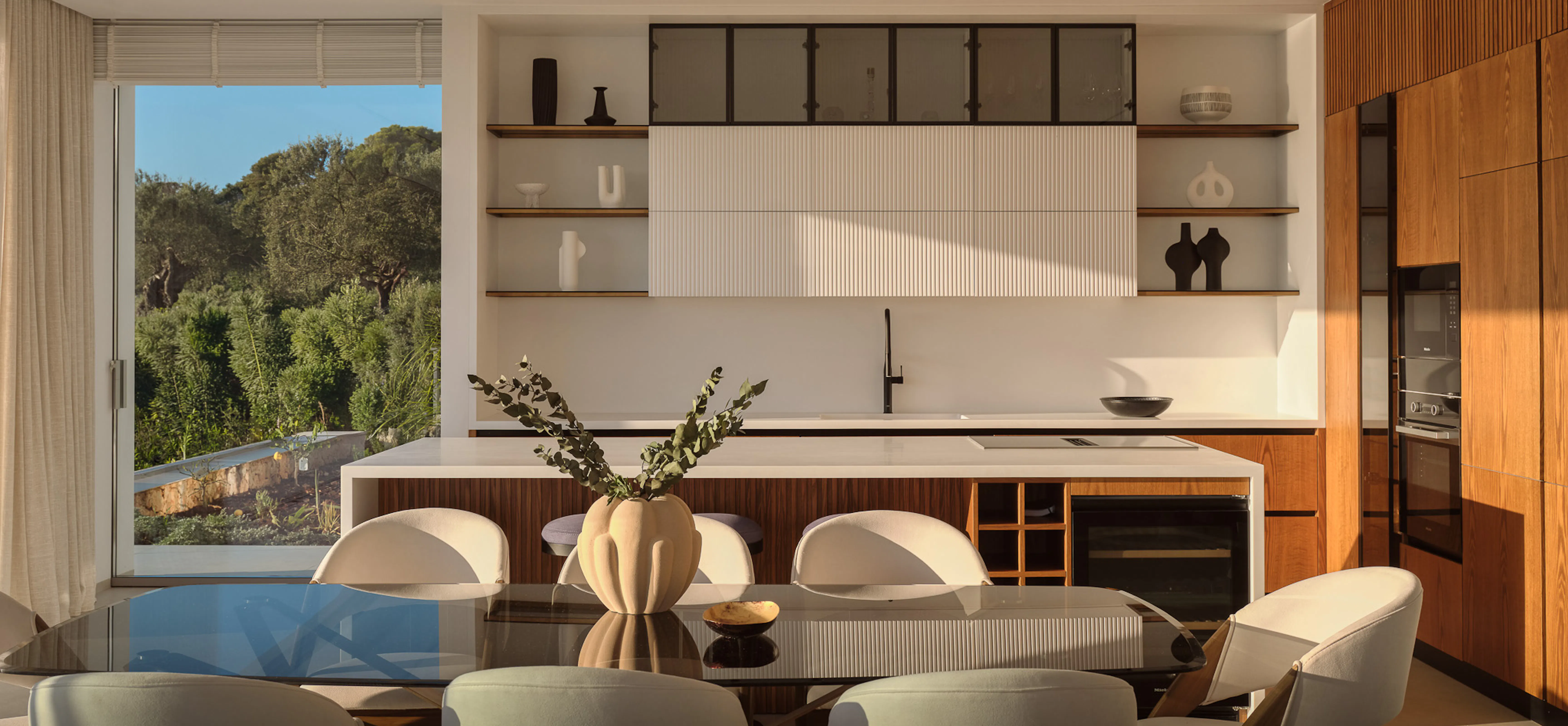 Sophisticated kitchen-dining room with sage green upholstered chairs around a dark oval table, warm walnut cabinetry, fluted white panels, and curated open shelving — refined yet inviting.