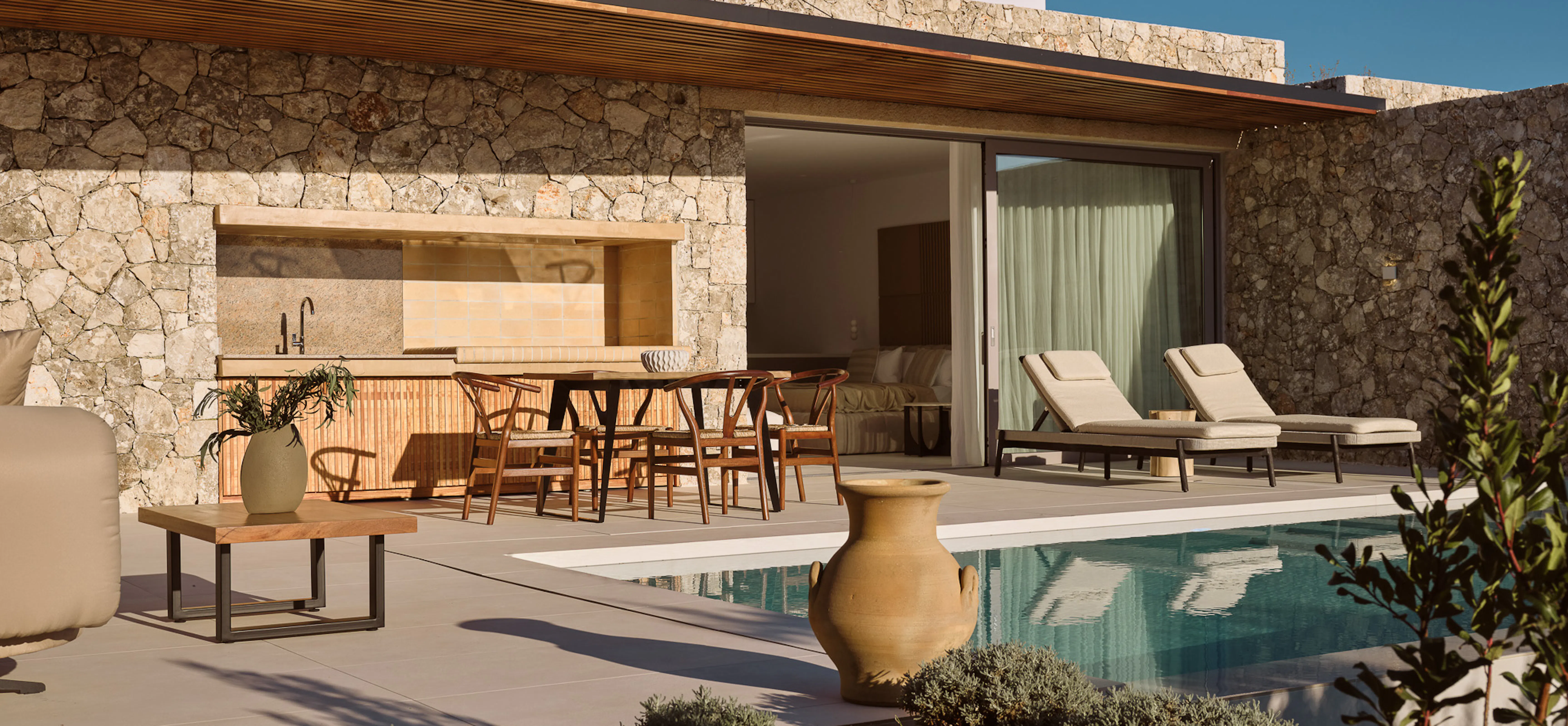 Villa exterior at golden hour showing a stone façade, outdoor kitchen, pool, and sun loungers surrounded by native Mediterranean planting — warm, tactile, and understated.