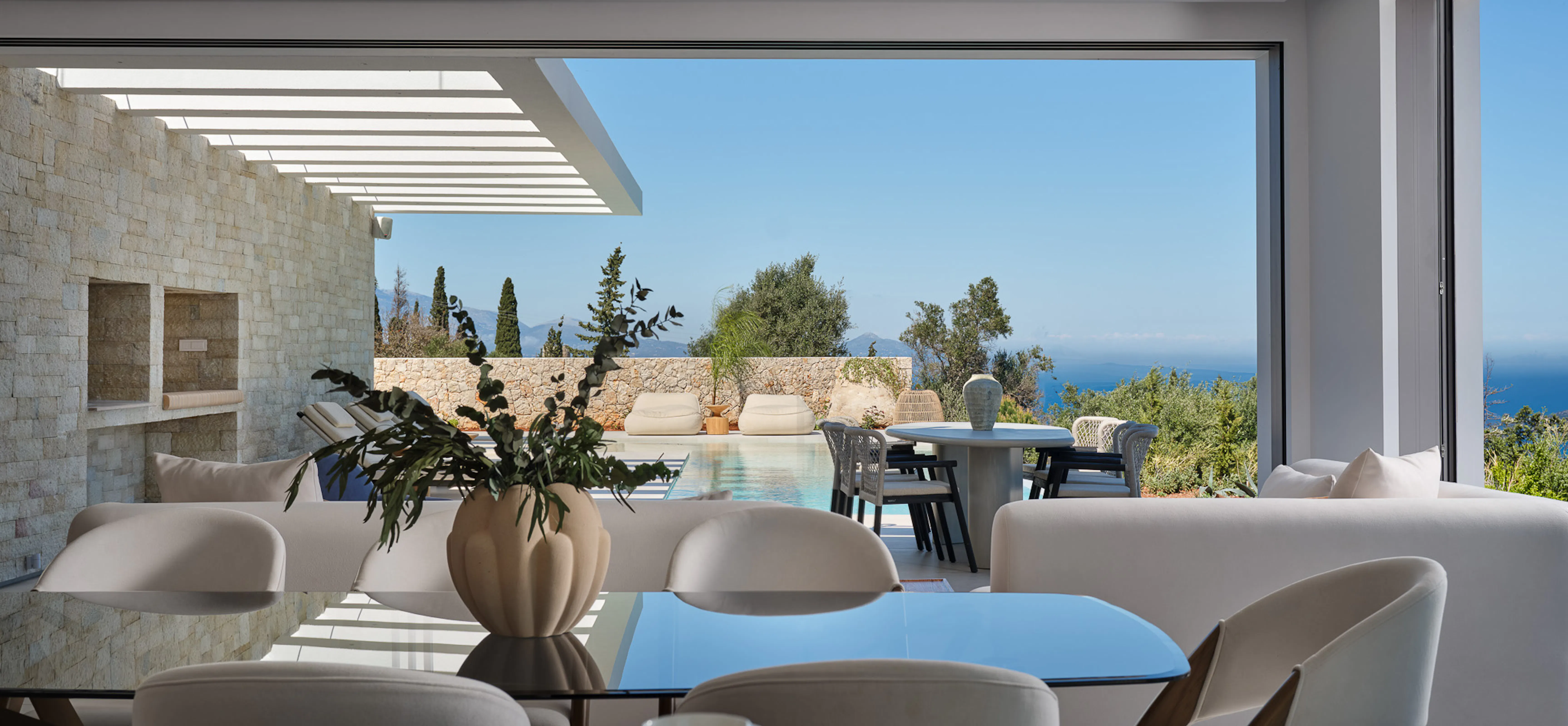 Sunlit interior dining area with a glass table and cream bouclé chairs, looking straight through to a pool terrace and the blue Ionian Sea — seamless inside-outside living.