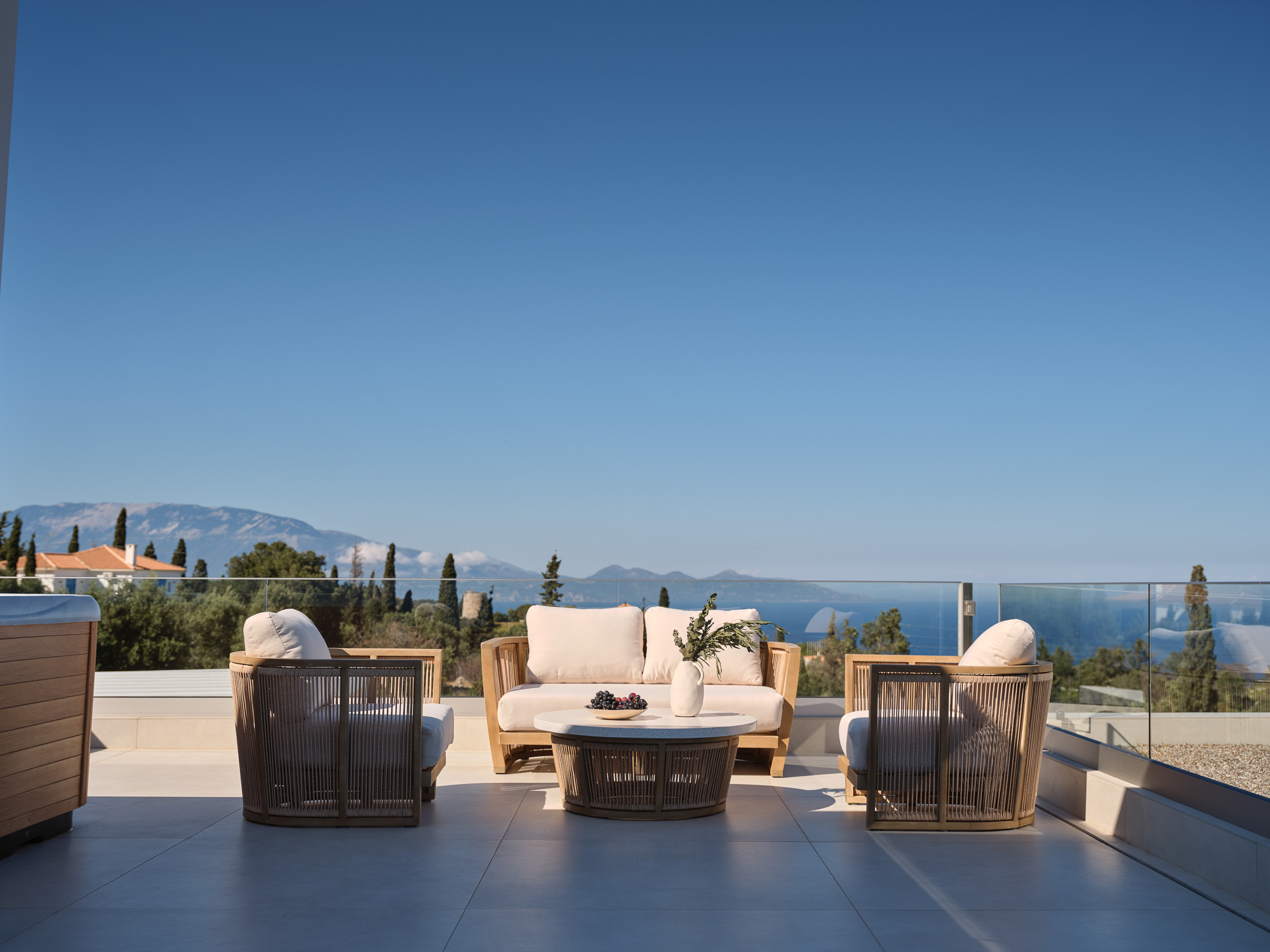 Rooftop terrace with teak and rope furniture, glass balustrade, and sweeping views over olive-covered hills to the blue sea below — open, breezy, and quietly sophisticated.