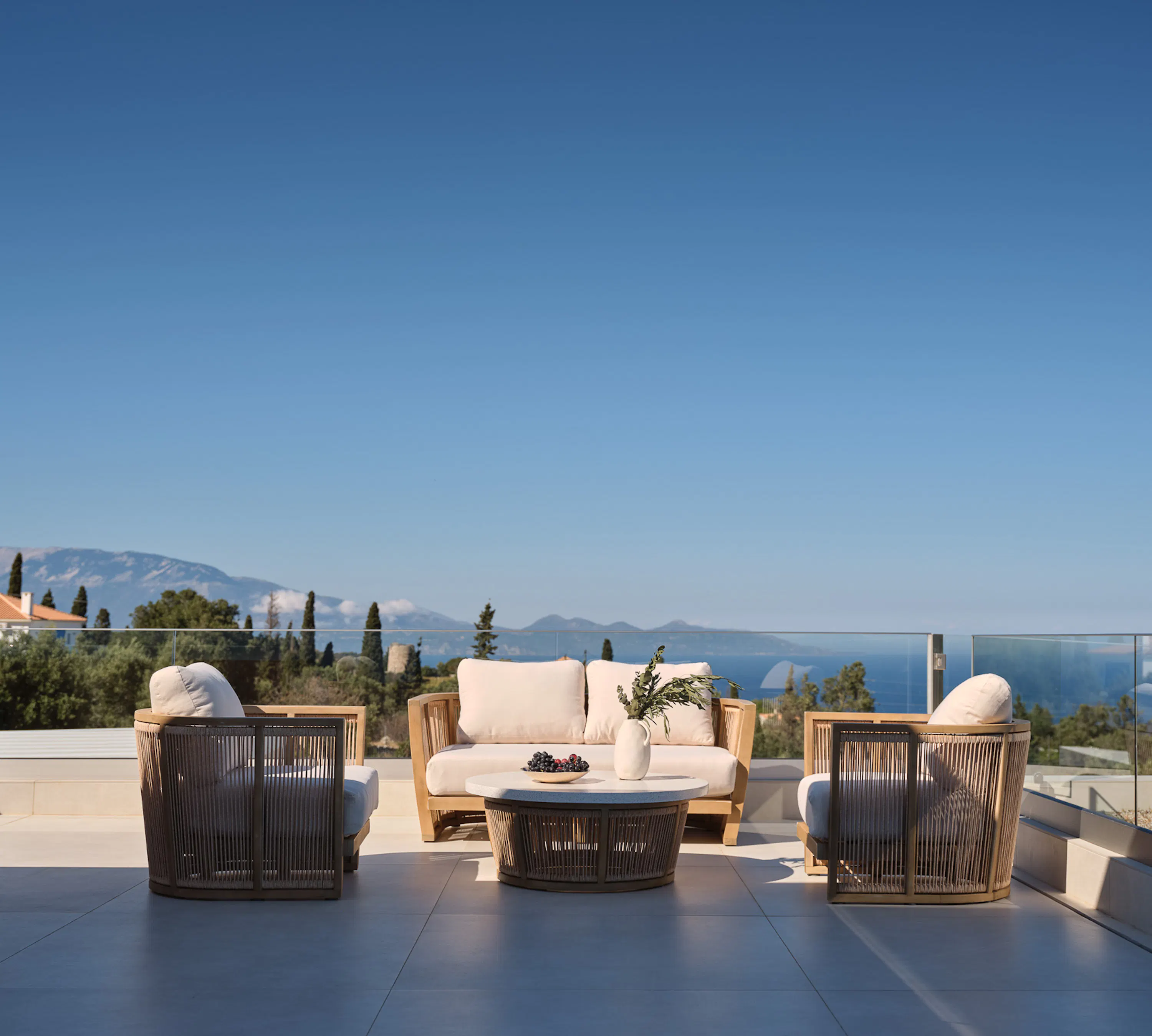 Rooftop terrace with teak and rope furniture, glass balustrade, and sweeping views over olive-covered hills to the blue sea below — open, breezy, and quietly sophisticated.