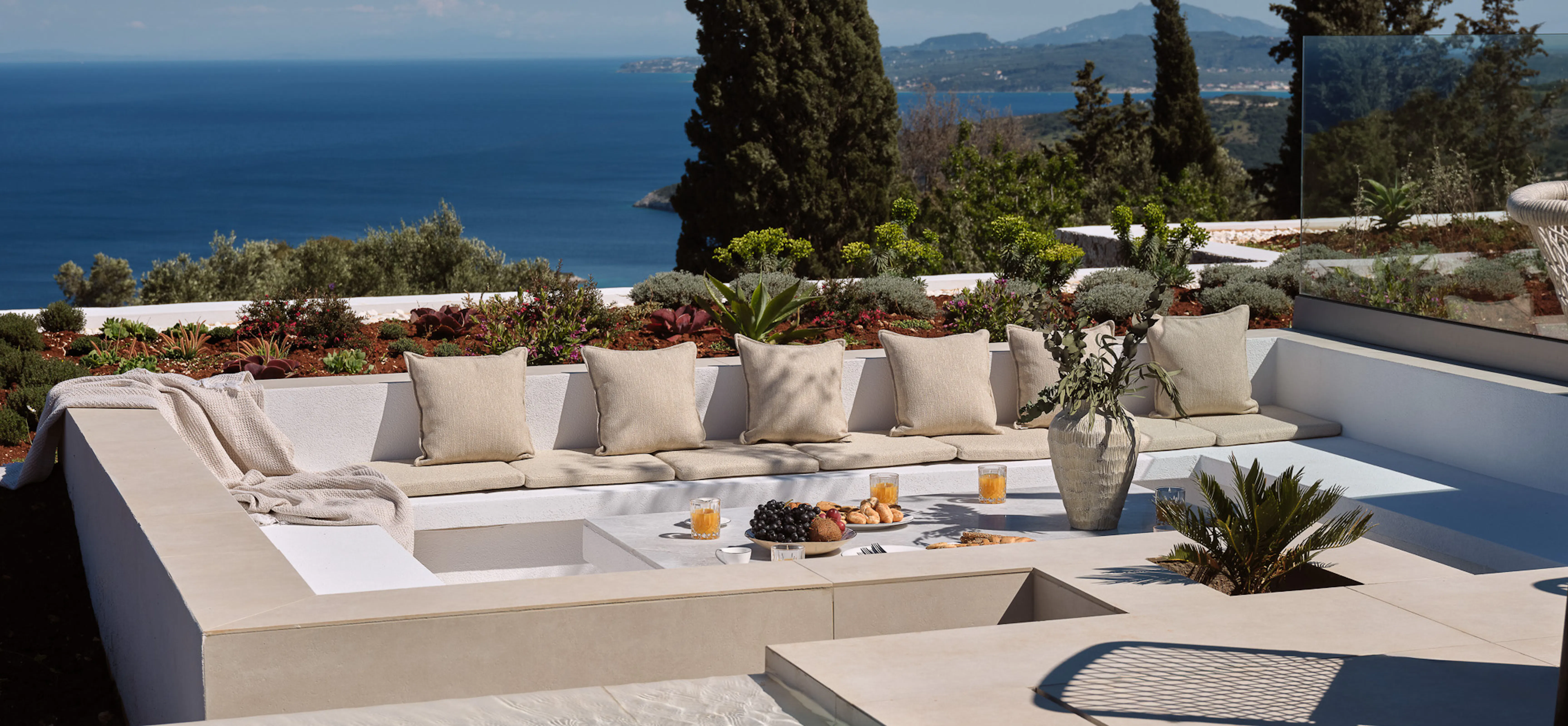 Sunken outdoor lounge with cream cushioned seating beside an infinity pool, overlooking a shimmering Ionian Sea panorama framed by cypress trees — relaxed Ionian luxury.