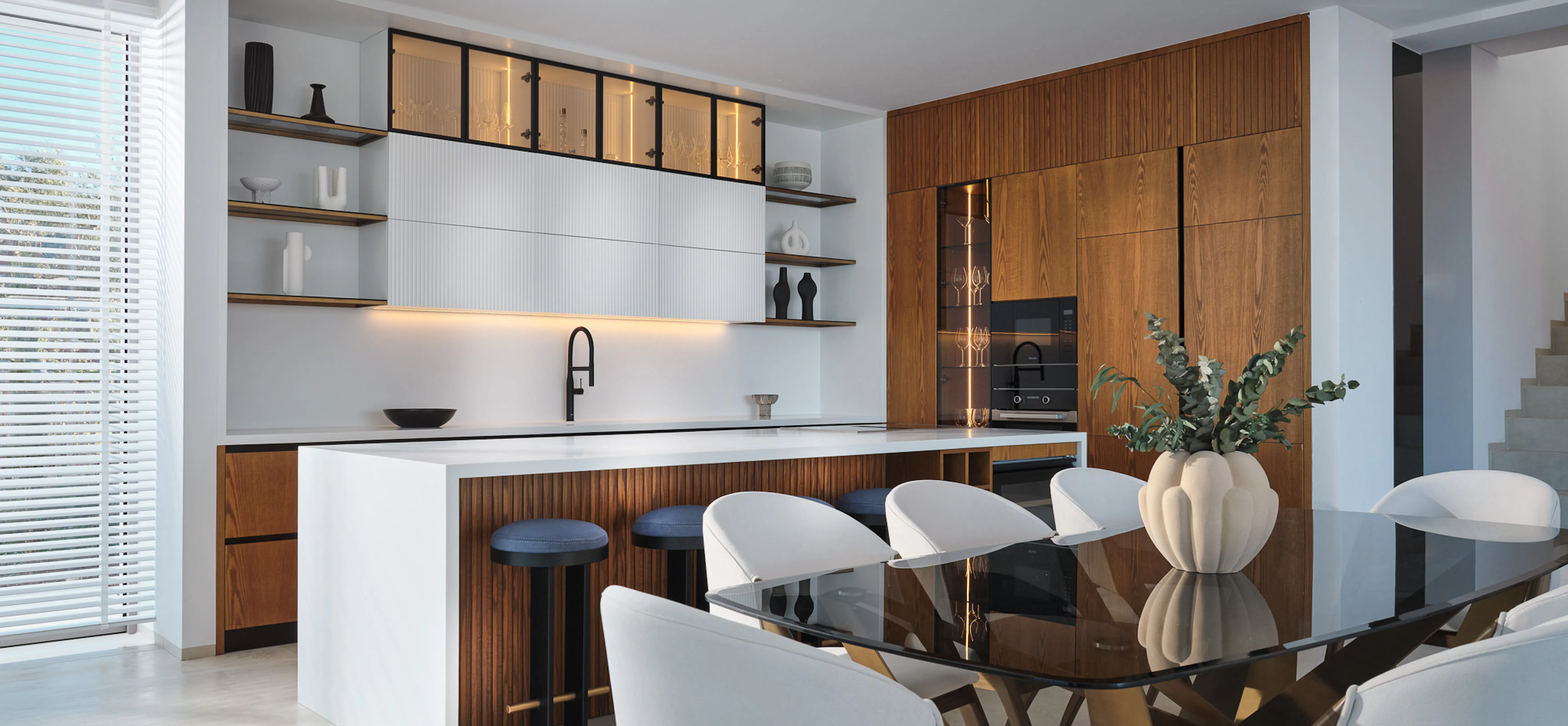 Stylish open-concept dining and kitchen area with white chairs, a black glass dining table, wood cabinetry, minimalist shelving, and bright natural light.
