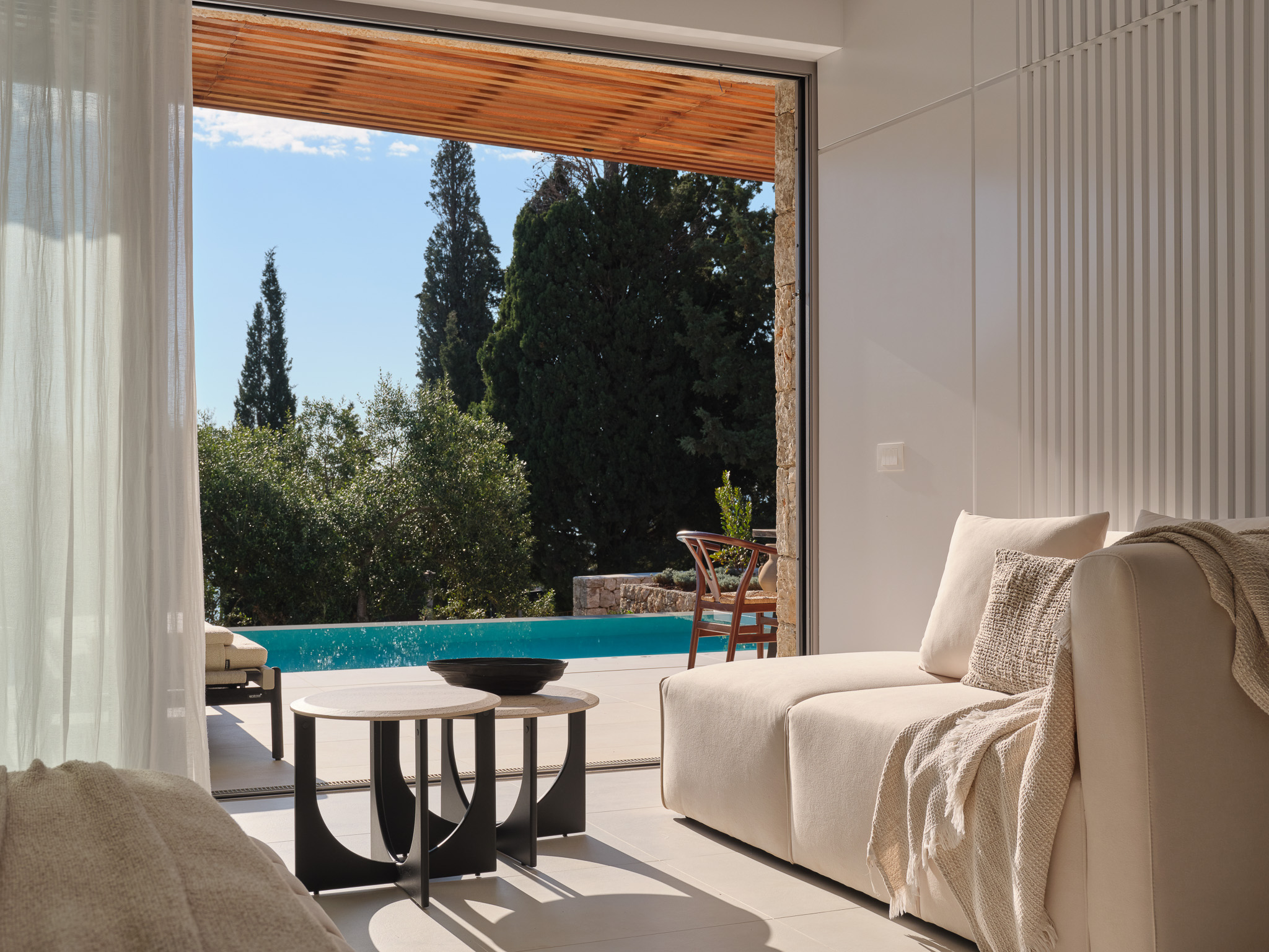 A bright, cloud-white sitting area with a deep, cushioned sofa draped in a textured throw, facing floor-to-ceiling glass that slides away entirely. Beyond lies a private pool shimmering in sunlight, surrounded by ancient olive and cypress trees. The wood-slatted ceiling overhang and sheer curtains filter the Greek afternoon light. The atmosphere is effortlessly elegant — intimate, tranquil, and deeply connected to the landscape.