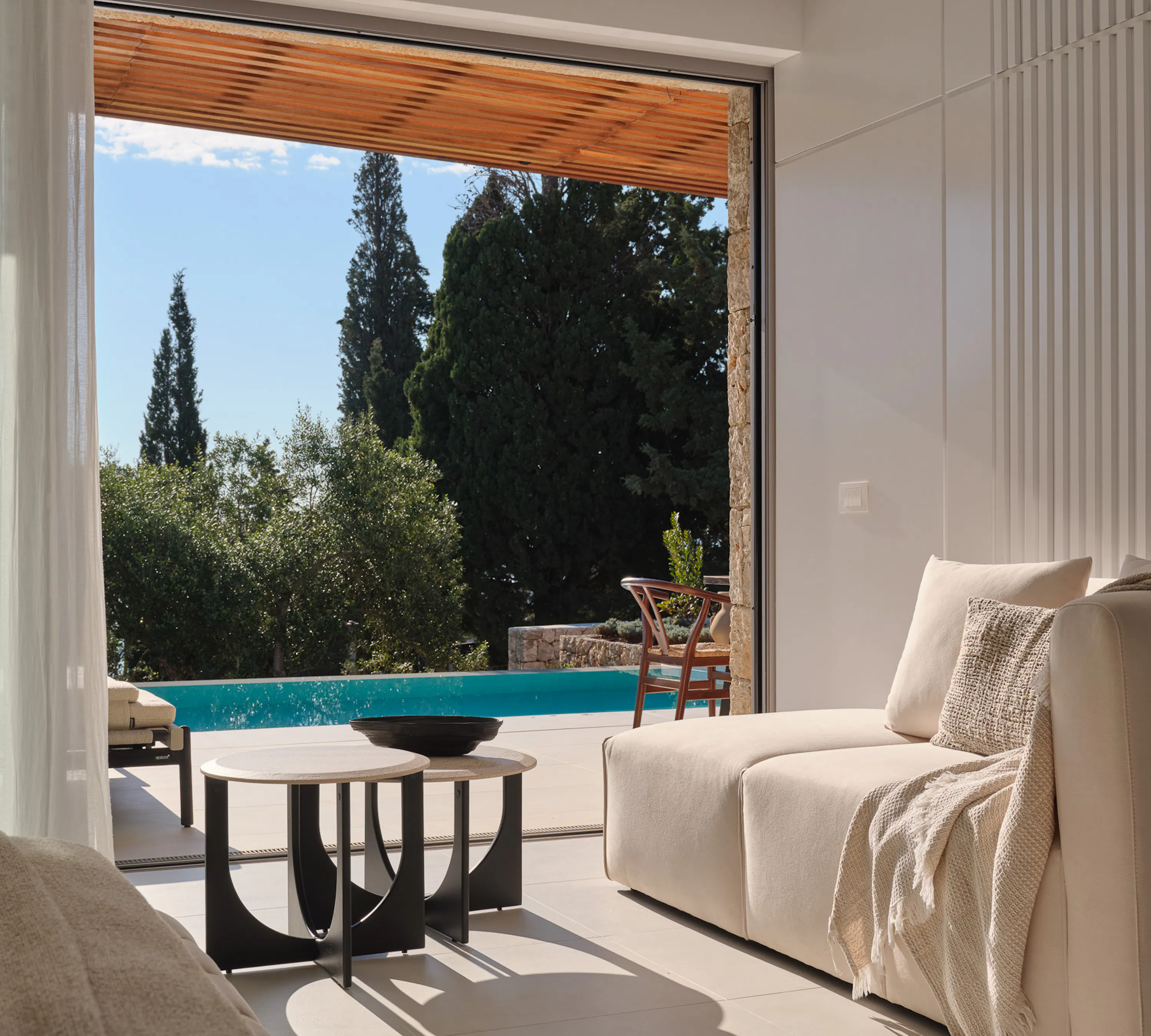 A bright, cloud-white sitting area with a deep, cushioned sofa draped in a textured throw, facing floor-to-ceiling glass that slides away entirely. Beyond lies a private pool shimmering in sunlight, surrounded by ancient olive and cypress trees. The wood-slatted ceiling overhang and sheer curtains filter the Greek afternoon light. The atmosphere is effortlessly elegant — intimate, tranquil, and deeply connected to the landscape.