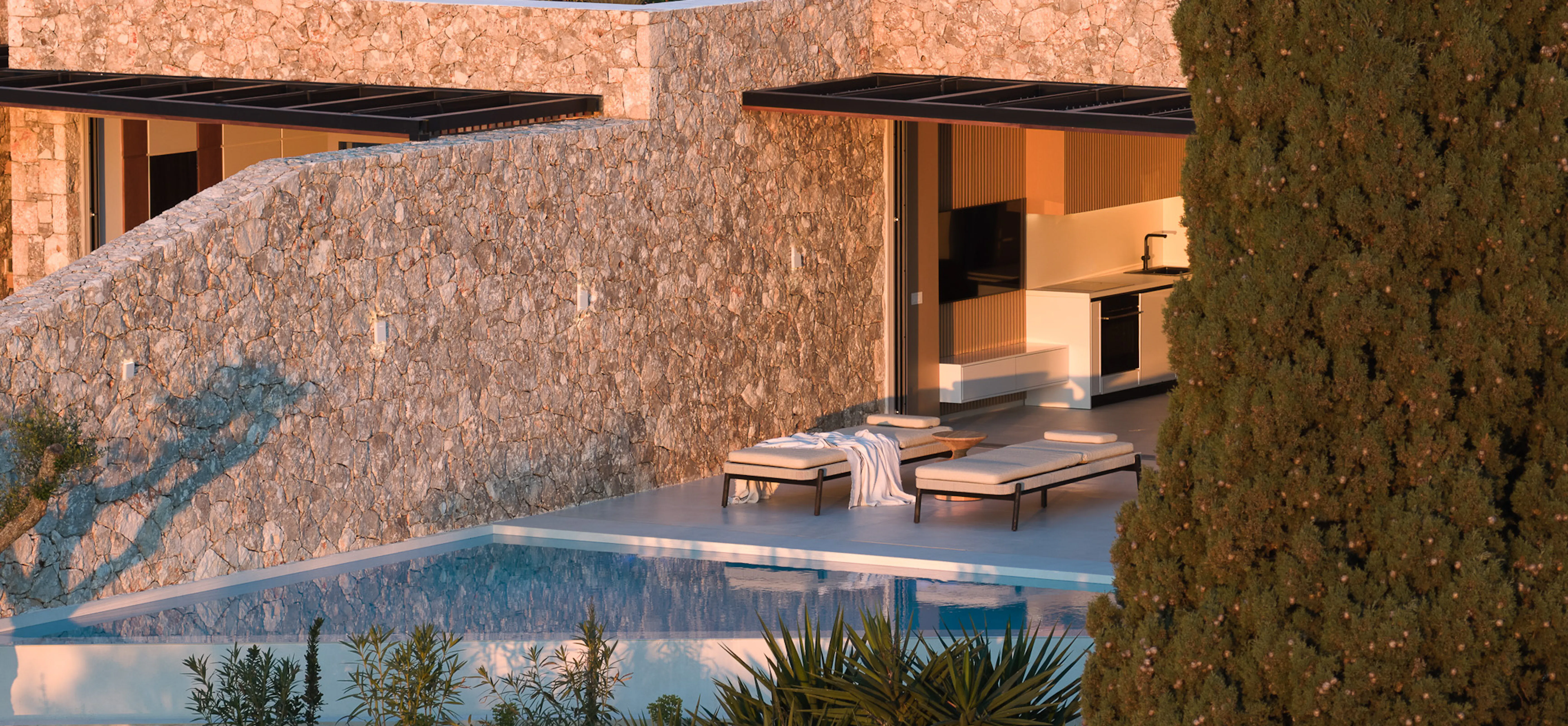 Golden evening light warms the ancient limestone walls of a low-lying villa carved into the hillside. Two slim sunbeds rest poolside beneath a wide overhang, with an outdoor kitchen glowing softly behind. A rooftop garden of succulents and wild Mediterranean plants crowns the structure. A tall cypress stands sentinel at the edge of frame. The atmosphere is primal yet polished — as if the building has always belonged to this rock and soil.