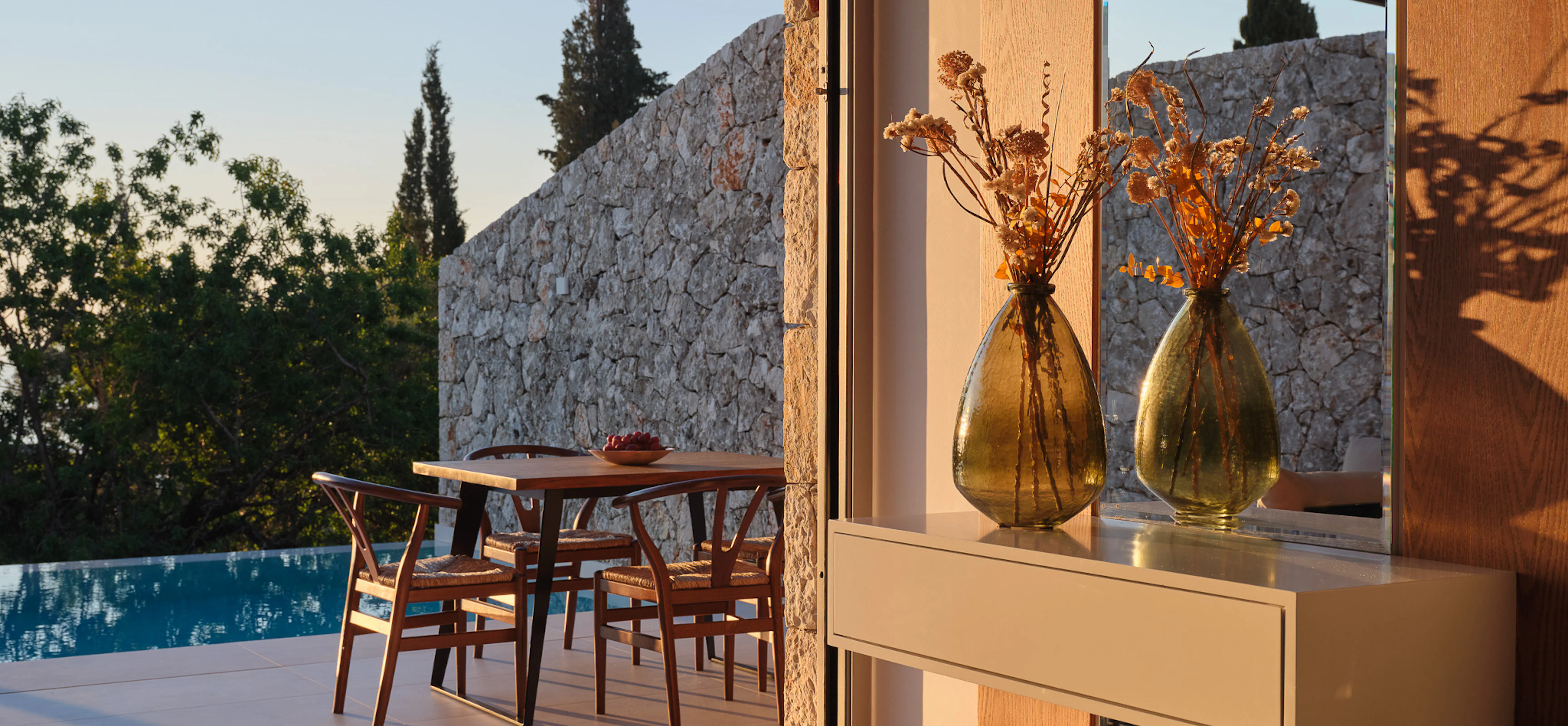 Warm amber light floods an open-plan terrace where a walnut dining table and wishbone chairs sit directly beside the pool. Two amber glass vases filled with dried botanical stems catch the last light on a floating sideboard. Stone walls and cypress trees form a natural enclosure beyond. The atmosphere is golden, unhurried, and deeply sensory — the smell of warm stone and evening air almost palpable.