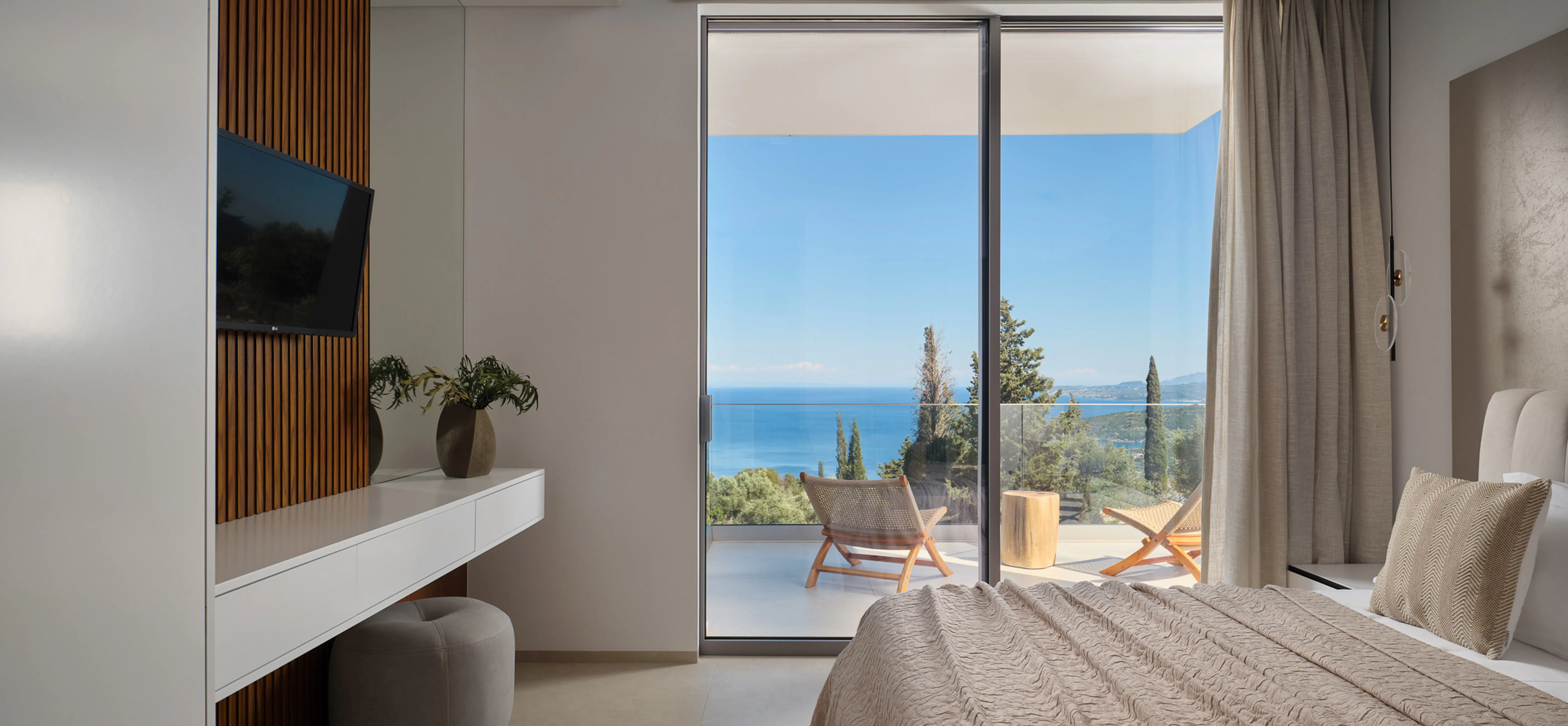 Crisp morning or afternoon light fills a minimal white bedroom. The sliding glass door frames a perfect rectangle of Ionian blue, cypress trees, and a balcony with wooden loungers. Warm timber slats, a floating desk, and a gold-footed stool complete the picture. The atmosphere is luminous and expansive — the sea feels like a living painting hung on the wall.