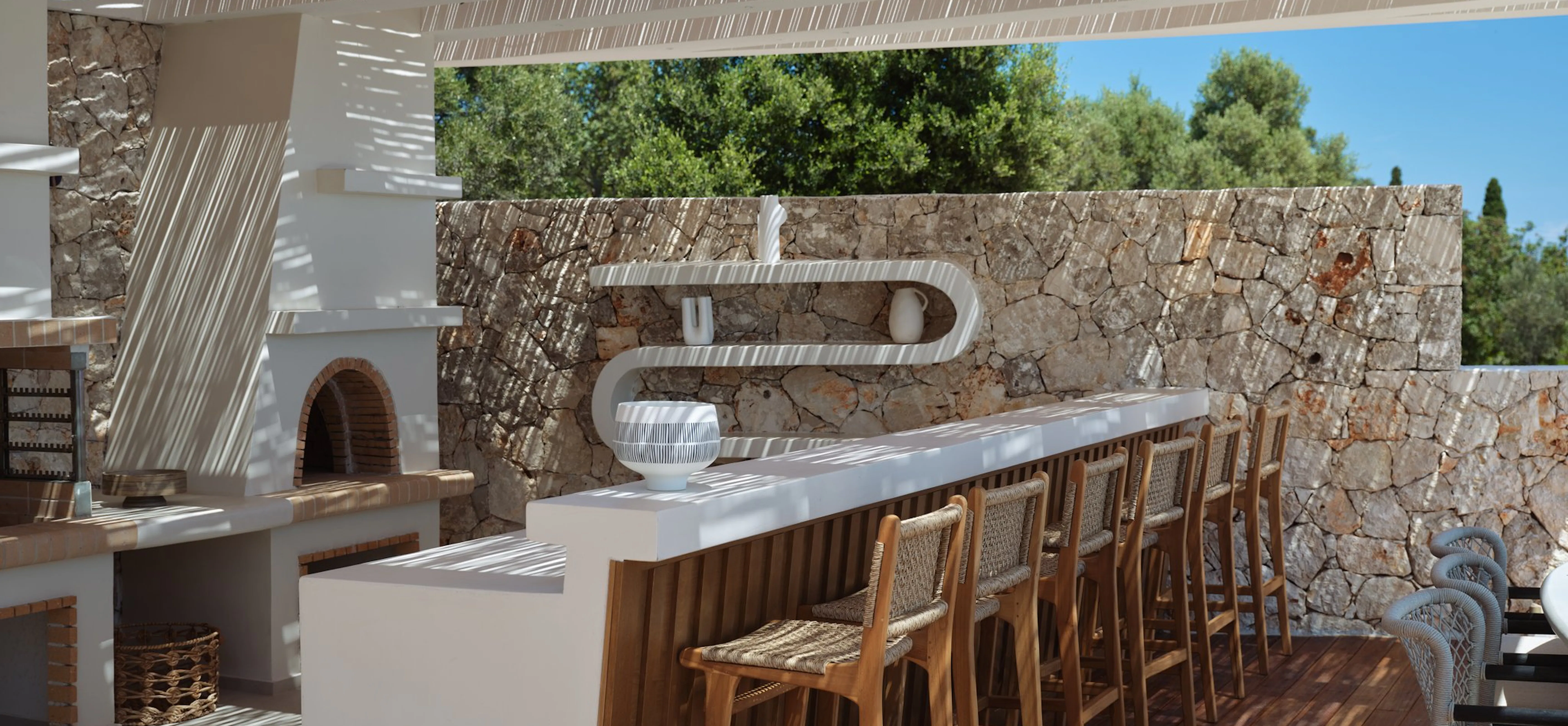 Rustic outdoor kitchen and bar area featuring a stone wall, wood-fired pizza oven, long marble countertop, and wooden bar stools beneath a slatted pergola.