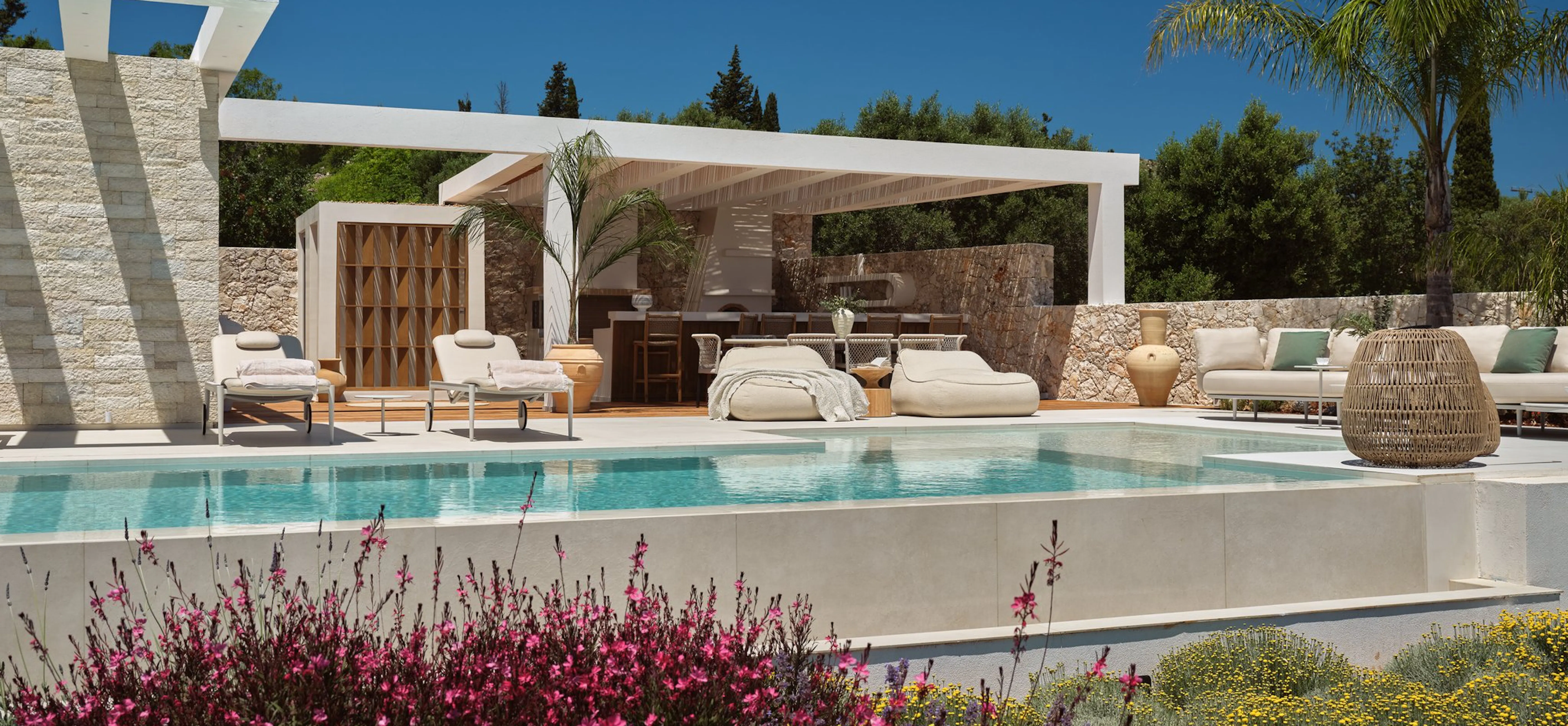 Luxury outdoor pool area with a white pergola, rattan lounge furniture, and lush pink and yellow flowering plants in the foreground under a bright blue sky.
