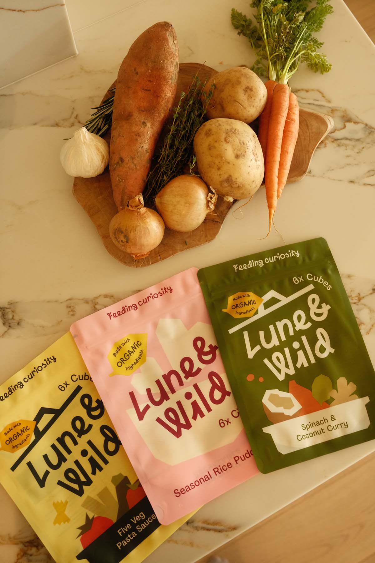 A collection of root vegetables including sweet potato, potatoes, onions, and carrots arranged on a marble surface, with three Luna & Wild product pouches in yellow, pink, and green displayed in front.