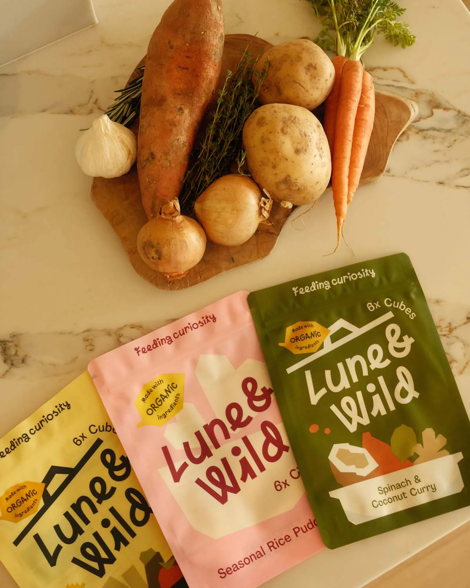 A collection of root vegetables including sweet potato, potatoes, onions, and carrots arranged on a marble surface, with three Luna & Wild product pouches in yellow, pink, and green displayed in front.