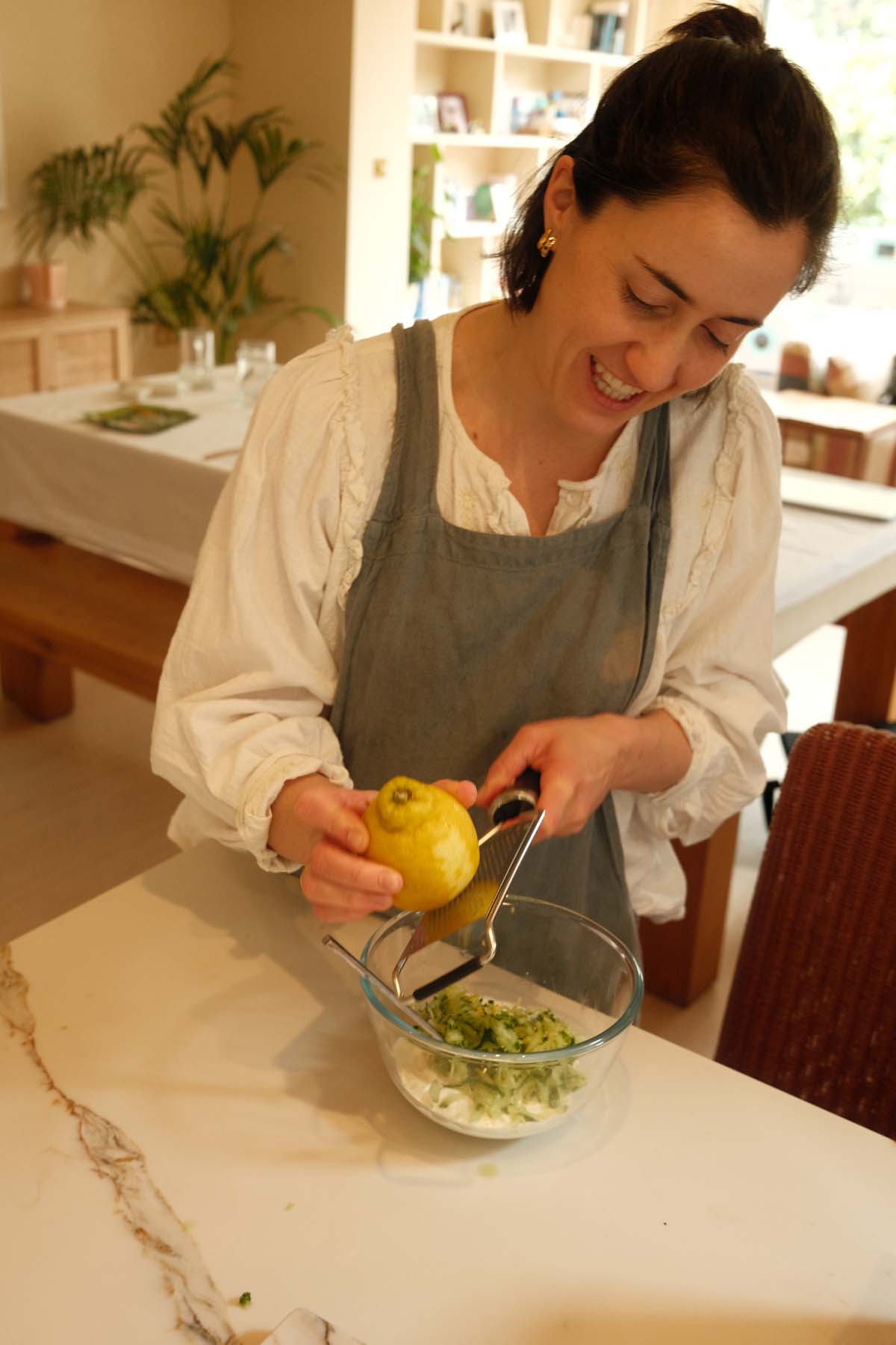 A woman in a linen apron squeezing a lemon into a glass bowl filled with chopped greens, standing at a white kitchen counter.