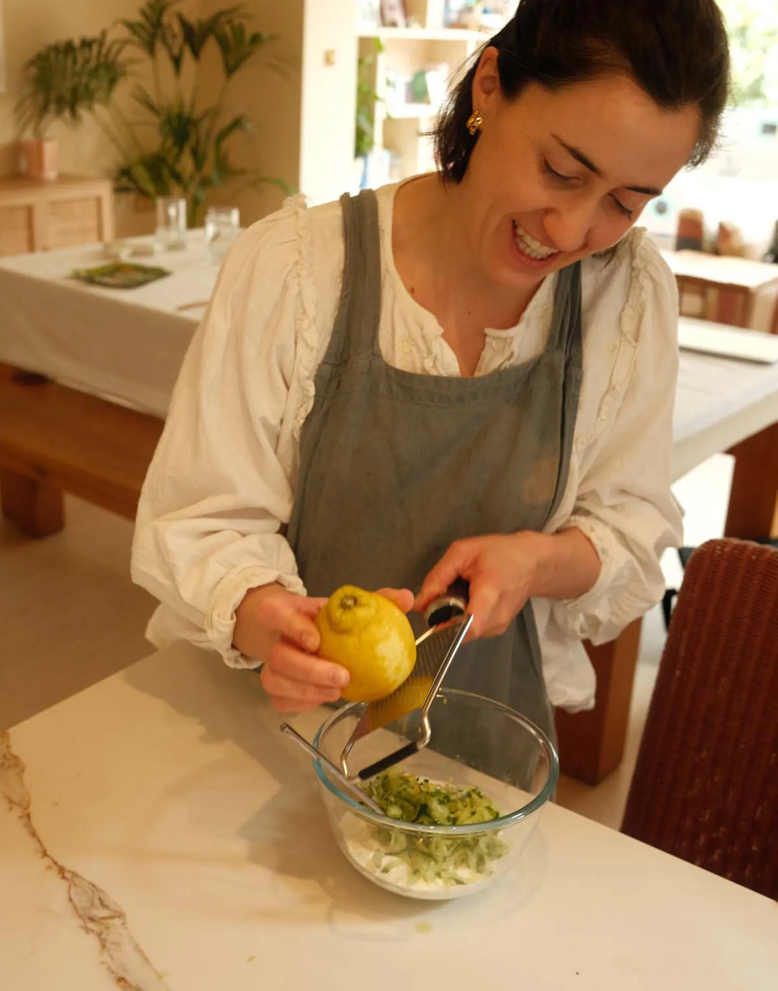 A woman in a linen apron squeezing a lemon into a glass bowl filled with chopped greens, standing at a white kitchen counter.