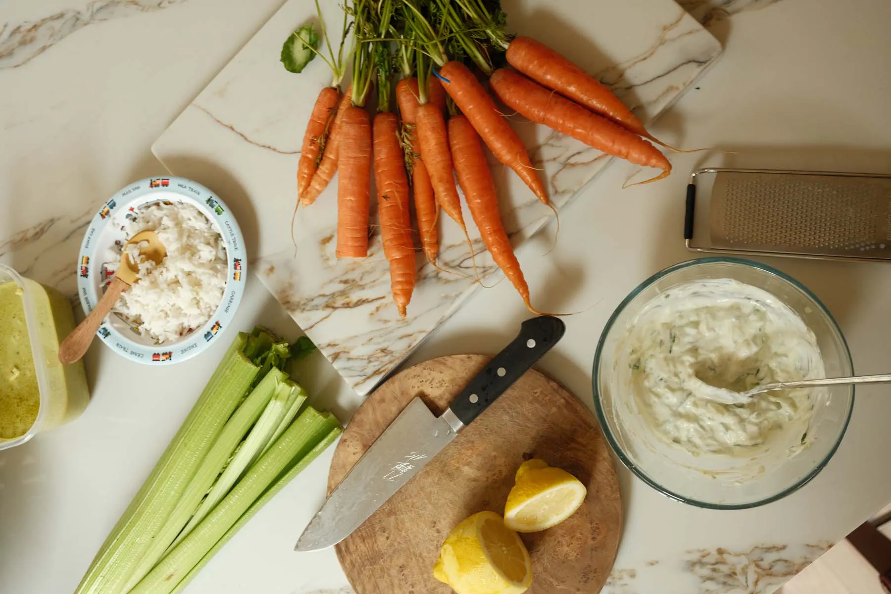 A flat lay of fresh ingredients on a wooden surface, including whole carrots, celery sticks, lemon wedges, and two small bowls of dip or spread, with a knife resting on a cutting board.