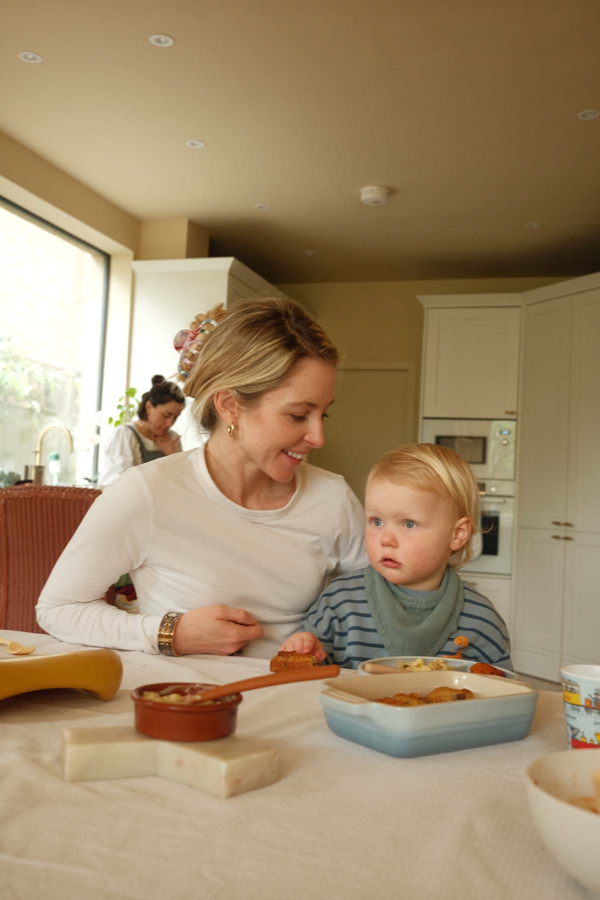 A woman sitting at a kitchen table with a young toddler on her lap, both looking at food on the table, with a jar and bowl visible nearby.