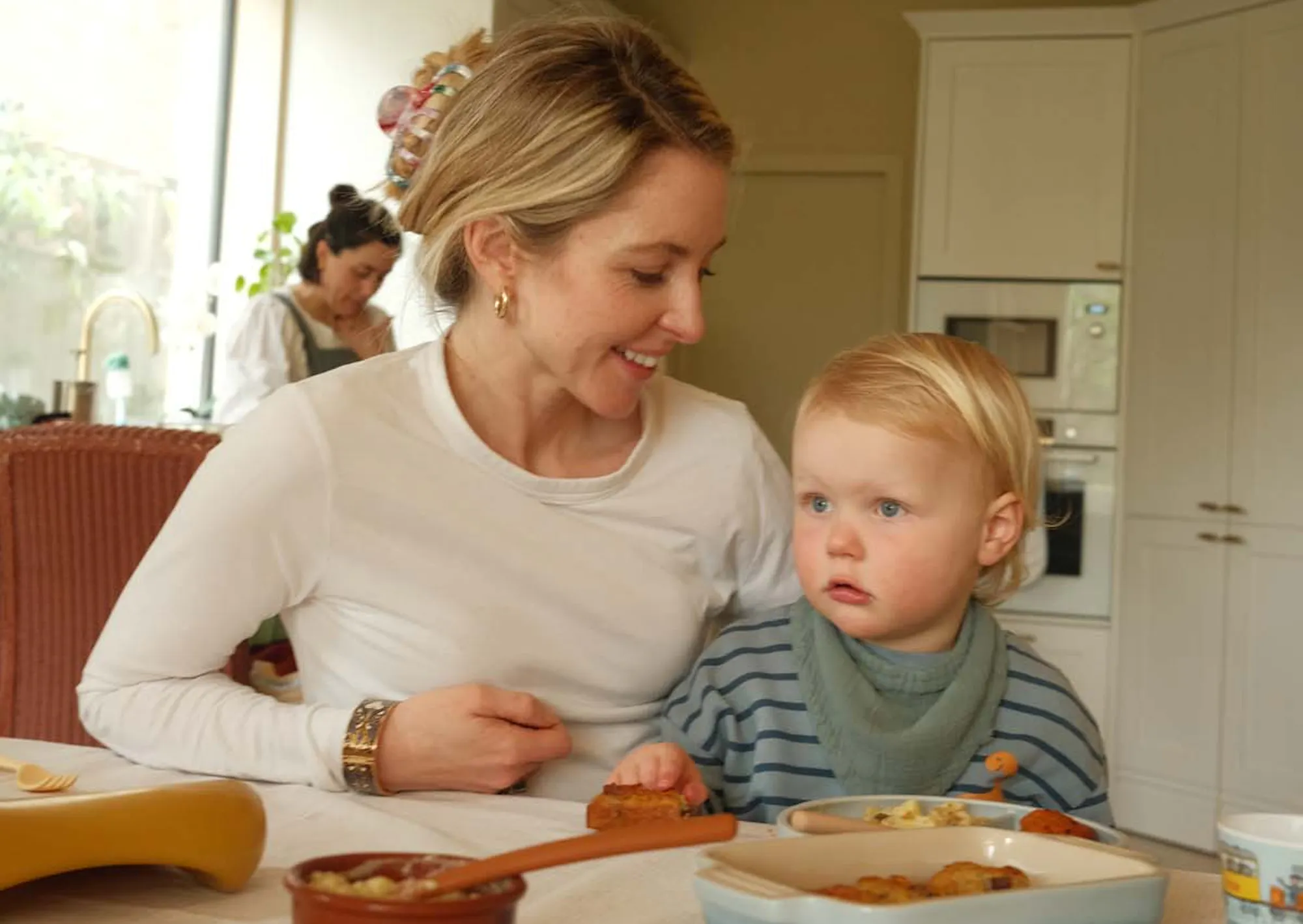 A woman sitting at a kitchen table with a young toddler on her lap, both looking at food on the table, with a jar and bowl visible nearby.