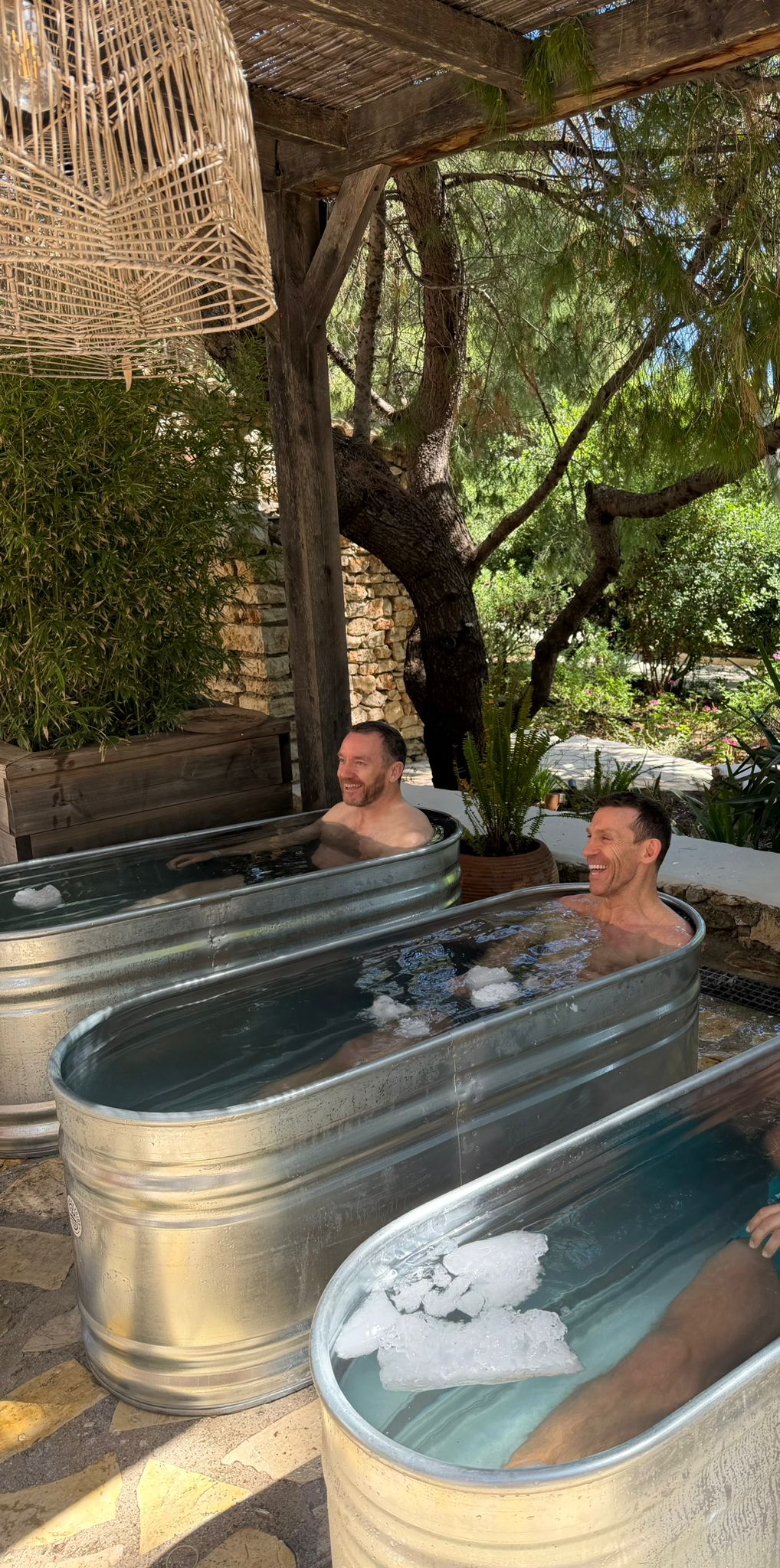 Two men sitting in separate galvanized metal ice bath tubs outdoors, surrounded by lush green trees and a woven rattan light fixture overhead. Both are smiling and appear relaxed in a rustic, garden-like wellness setting.
