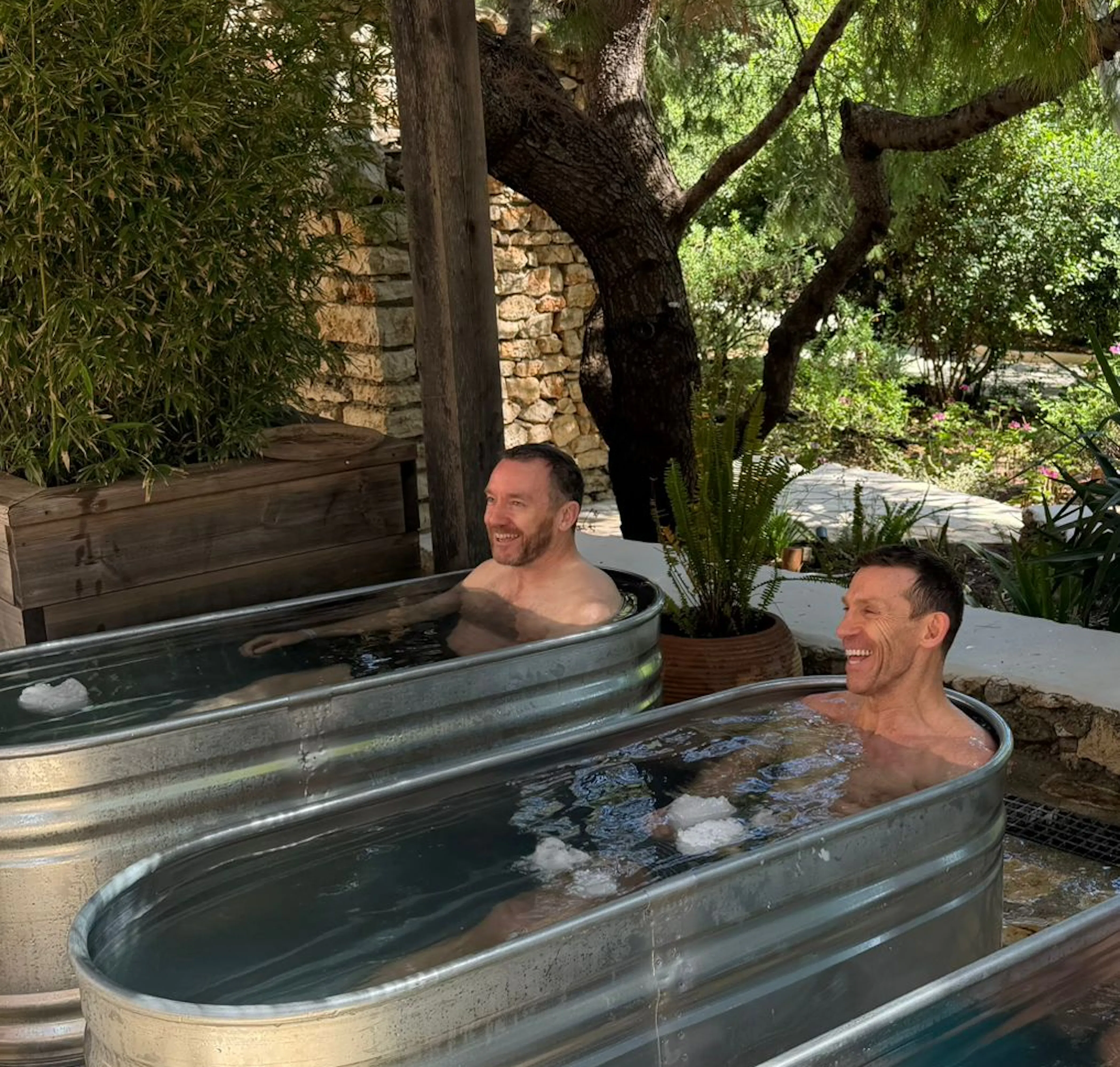Two men sitting in separate galvanized metal ice bath tubs outdoors, surrounded by lush green trees and a woven rattan light fixture overhead. Both are smiling and appear relaxed in a rustic, garden-like wellness setting.
