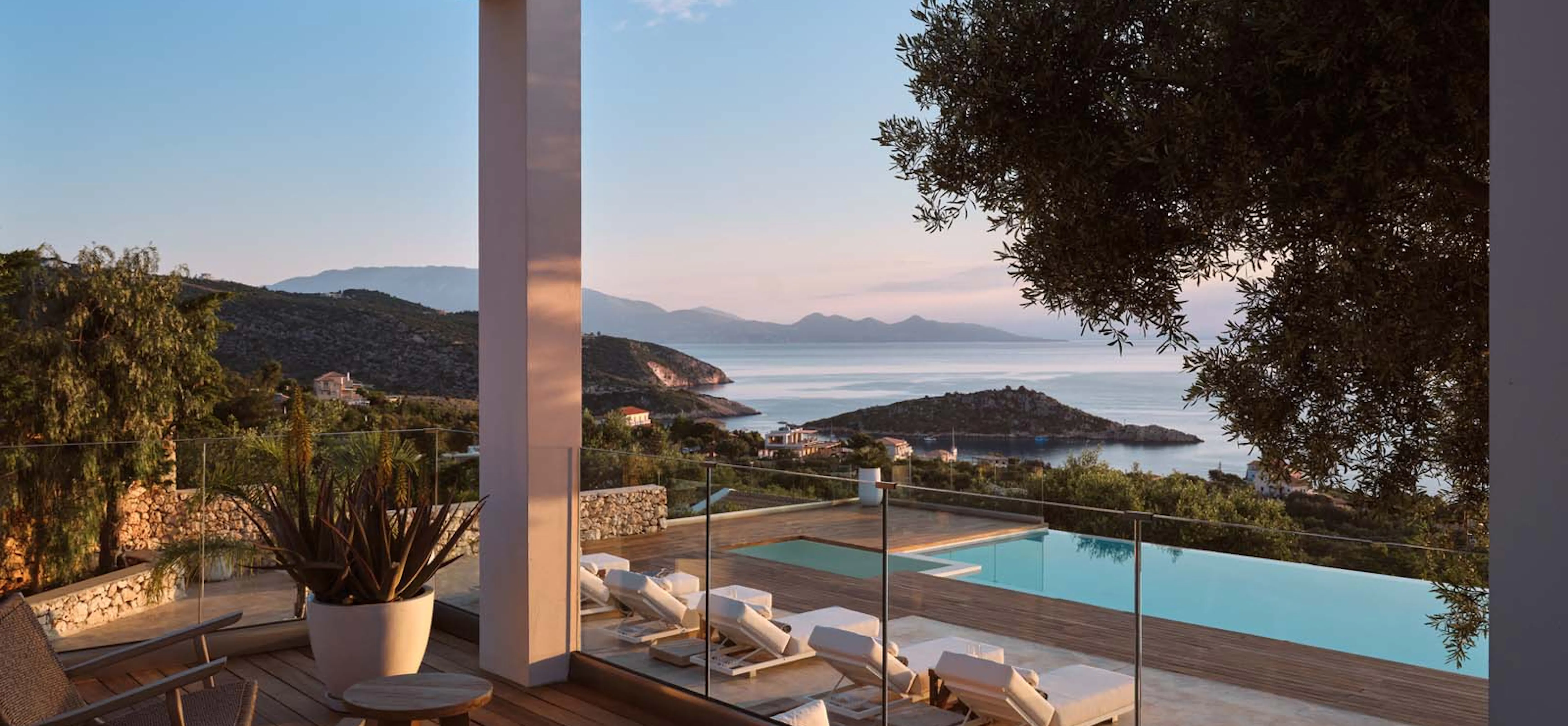 Elevated villa terrace at dusk with wicker chairs on a wood deck, overlooking a pool lined with sun loungers, a hillside village, and a calm bay with islands fading into a pastel evening sky.