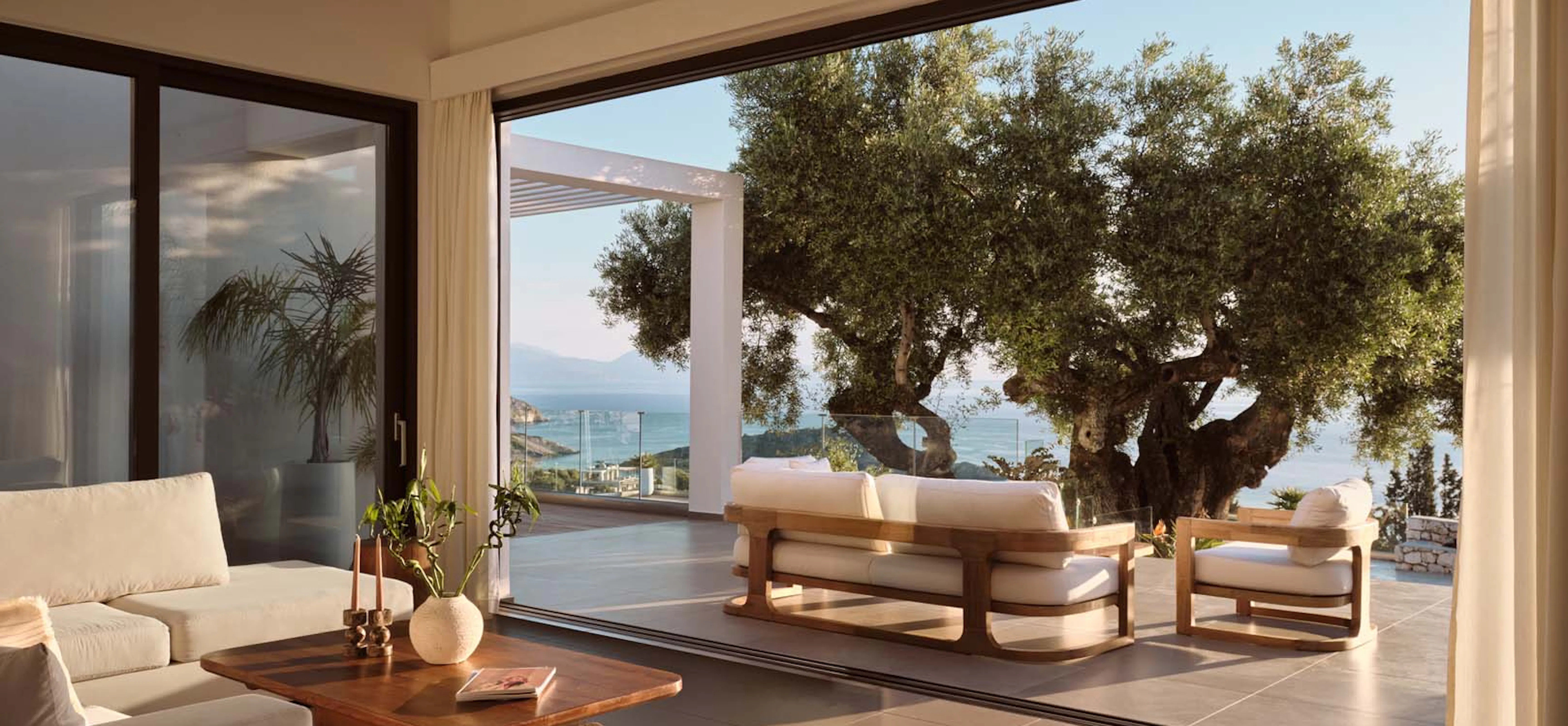 Bright villa living room with white sofas and a walnut coffee table, opening through floor-to-ceiling sliding glass doors onto an outdoor lounge area shaded by a large olive tree, with sea and mountain views.