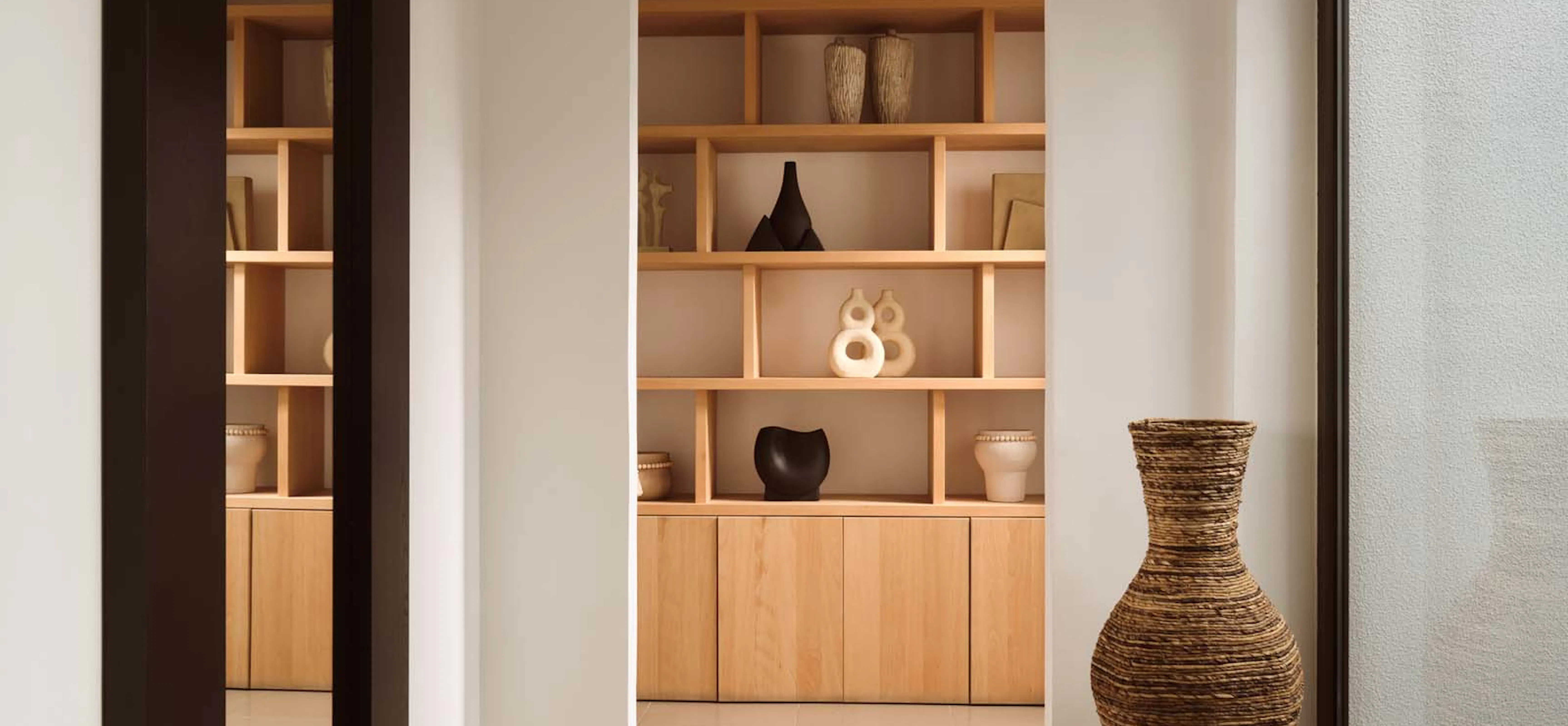 Minimalist villa hallway with a large floor mirror, a tall woven floor vase, and a built-in light wood shelving unit displaying sculptural ceramic objects in neutral tones.