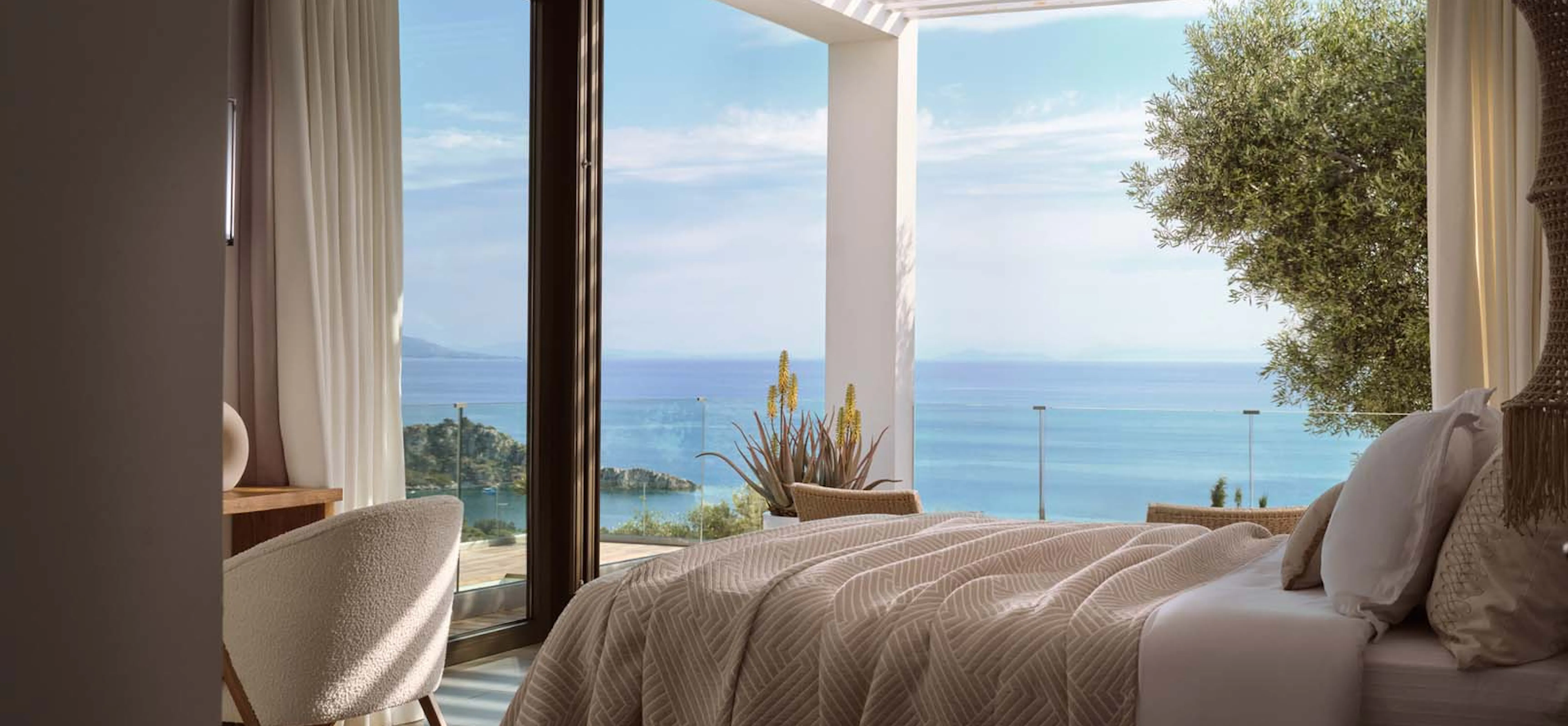 Bright, airy villa bedroom with a textured beige bedspread, a bouclé armchair, and floor-to-ceiling corner windows overlooking an olive tree, terrace pergola, and vivid blue sea.