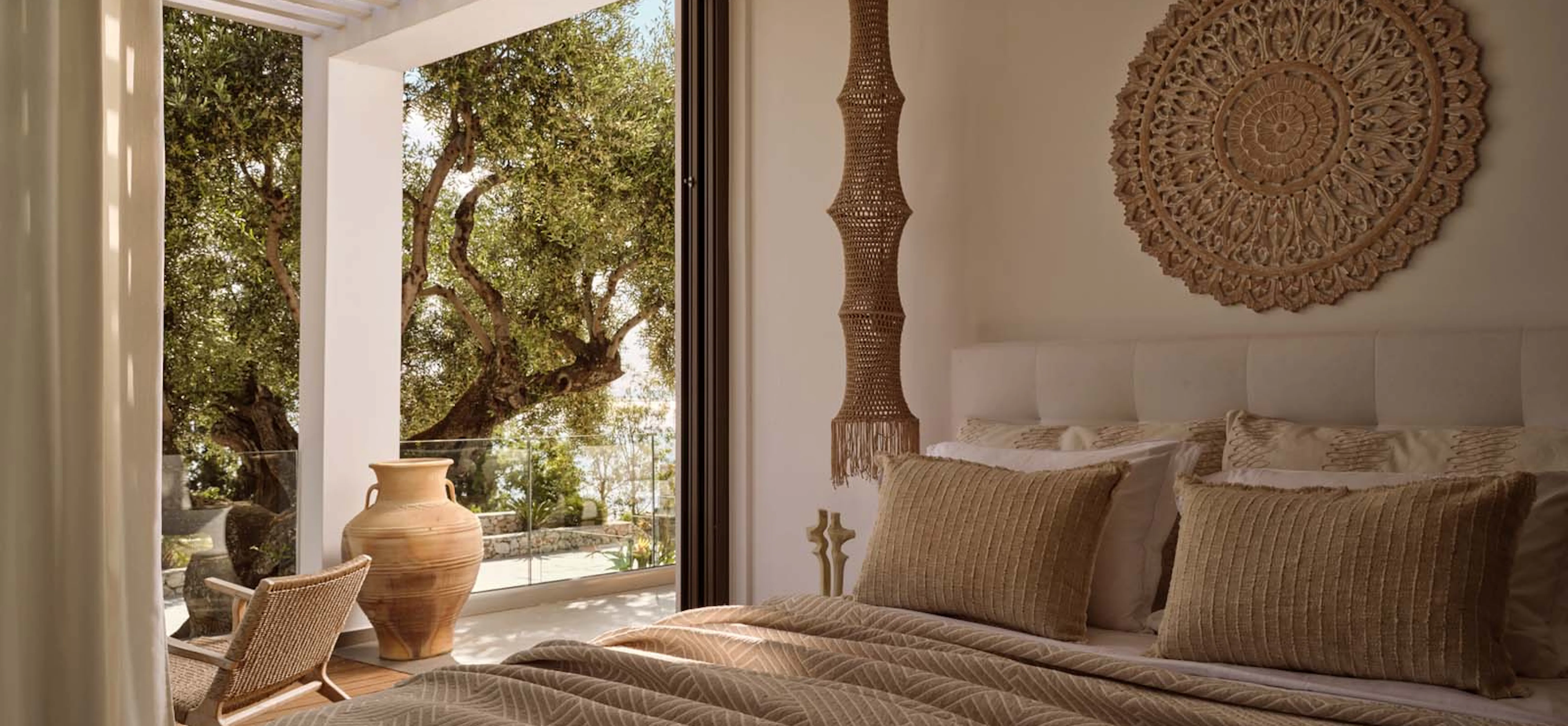 Villa bedroom with warm earth-toned bedding, a large carved mandala wall decoration, a tall woven macramé pendant light, and an open sliding door revealing an olive tree and terrace.
