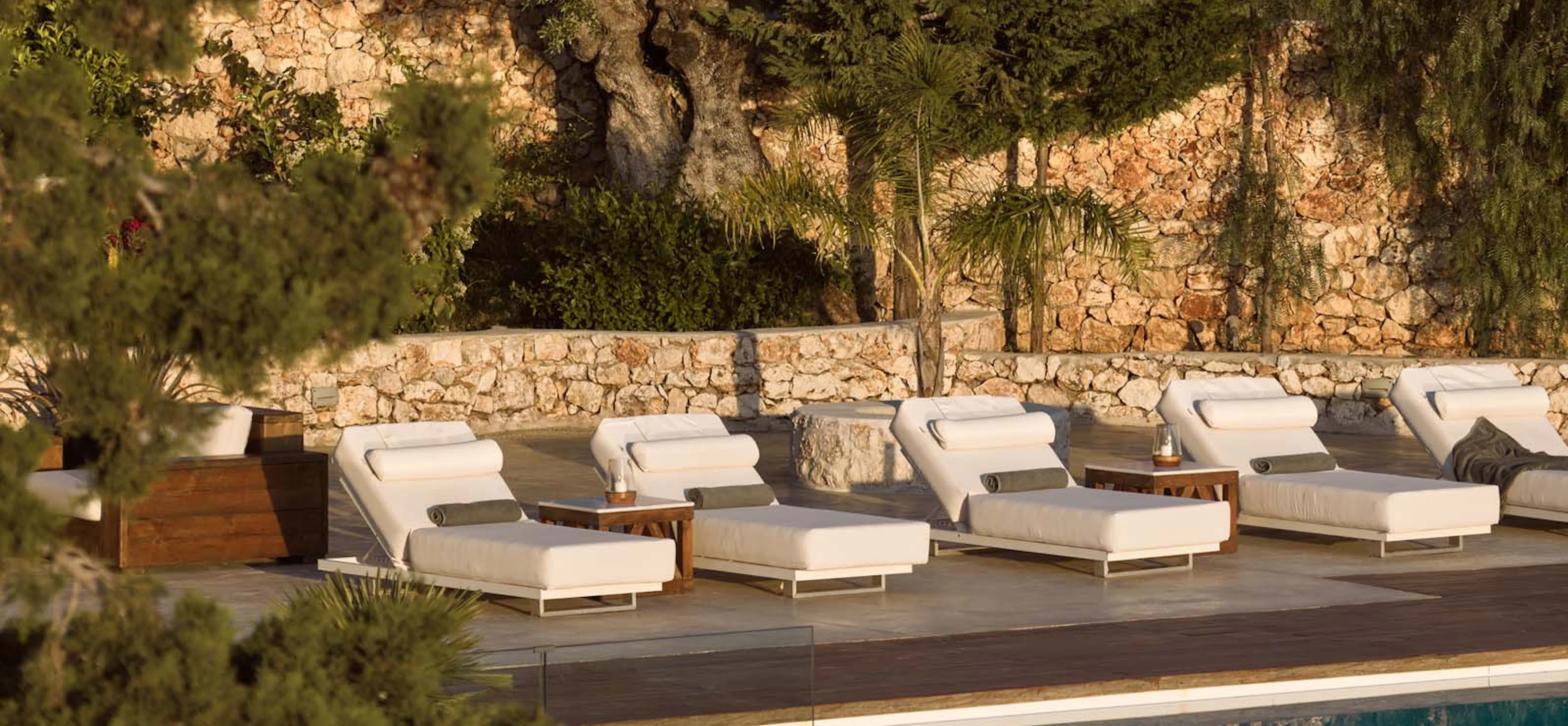 A row of white-cushioned sun loungers with rolled towels beside a pool, backed by a golden-lit stone wall and lush olive and cypress trees.