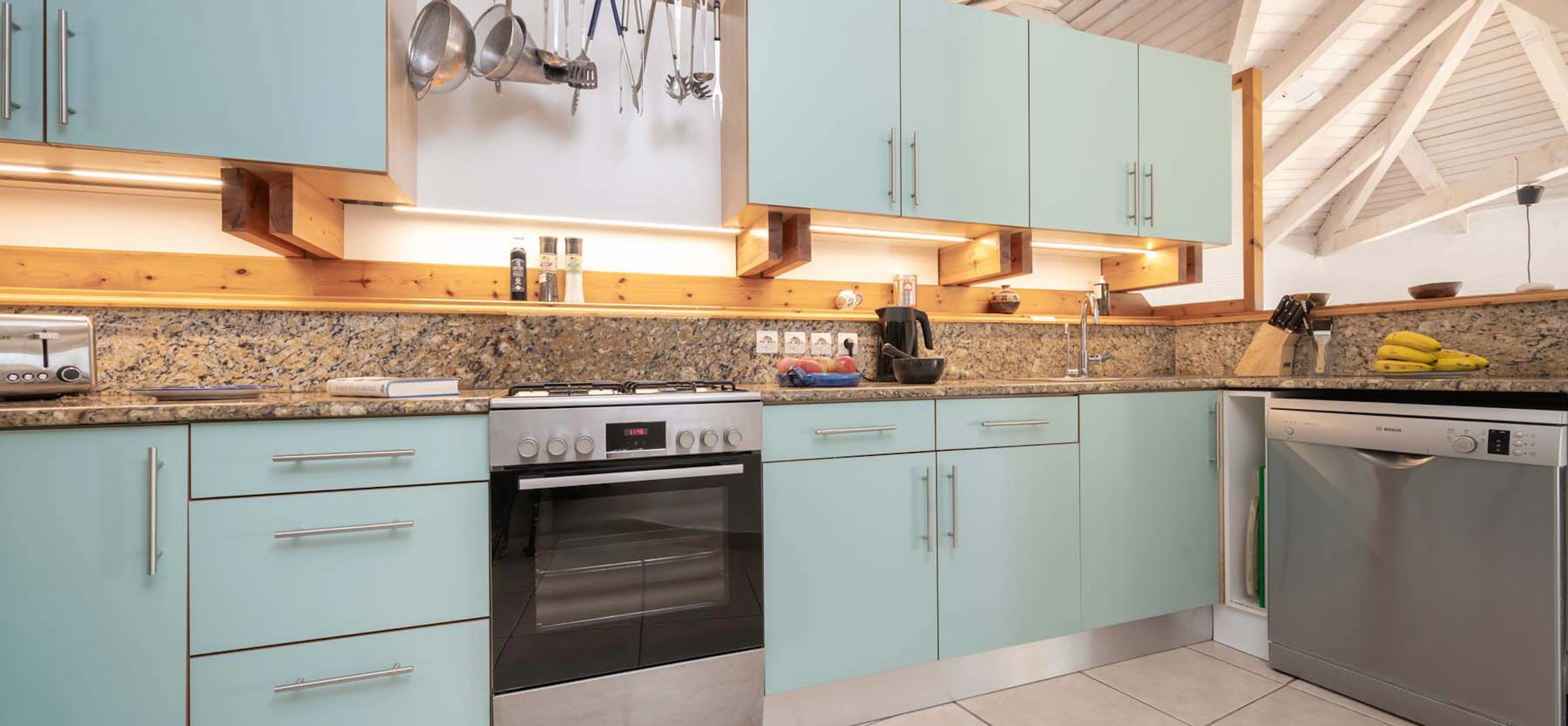 A well-equipped kitchen with pale duck-egg blue cabinetry, granite worktops with warm-lit pine shelving, a stainless steel gas oven, Bosch dishwasher, and hanging utensil rail beneath a whitewashed vaulted timber ceiling.