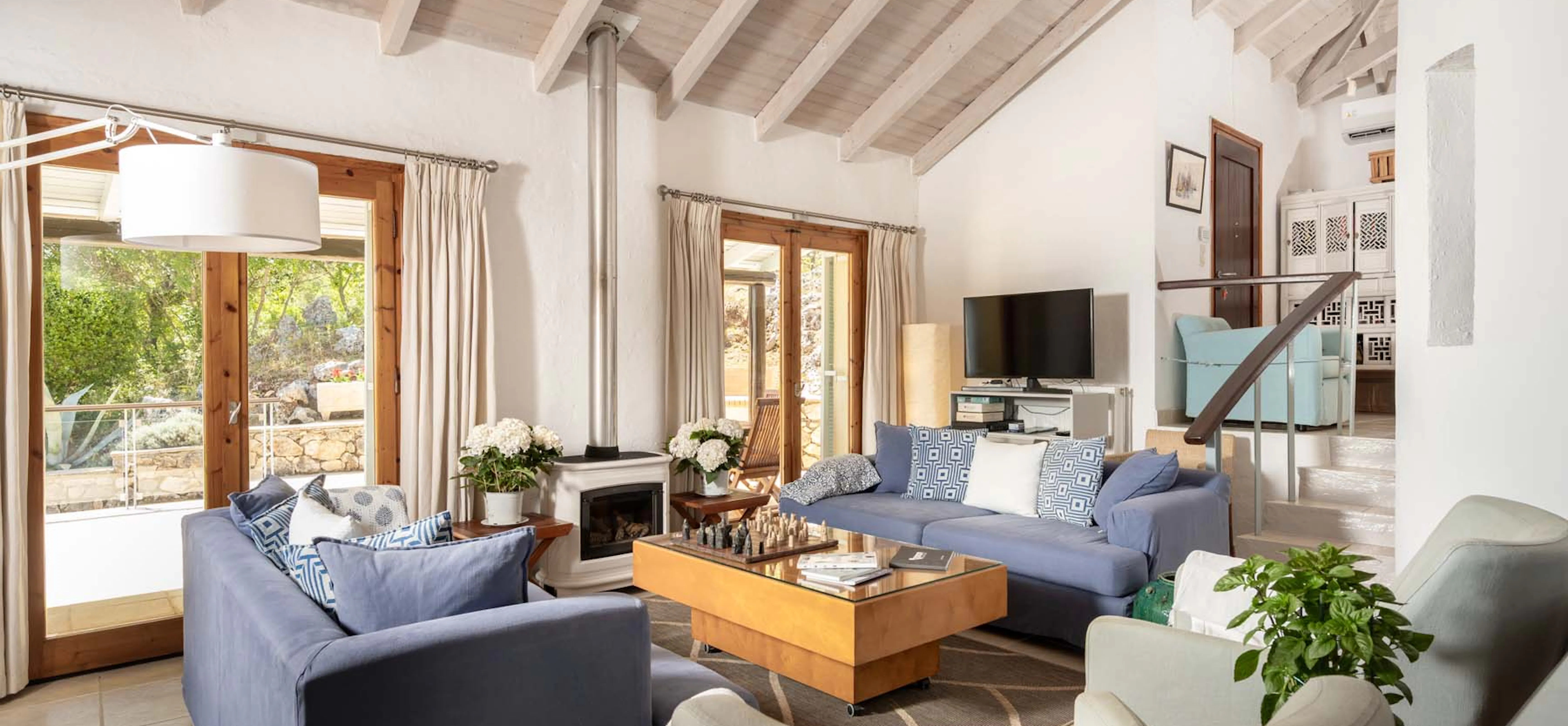A spacious living room with white vaulted beamed ceiling, blue linen sofas, a central wood-burning stove, glass coffee table with a chess set, and French doors opening to a garden terrace.