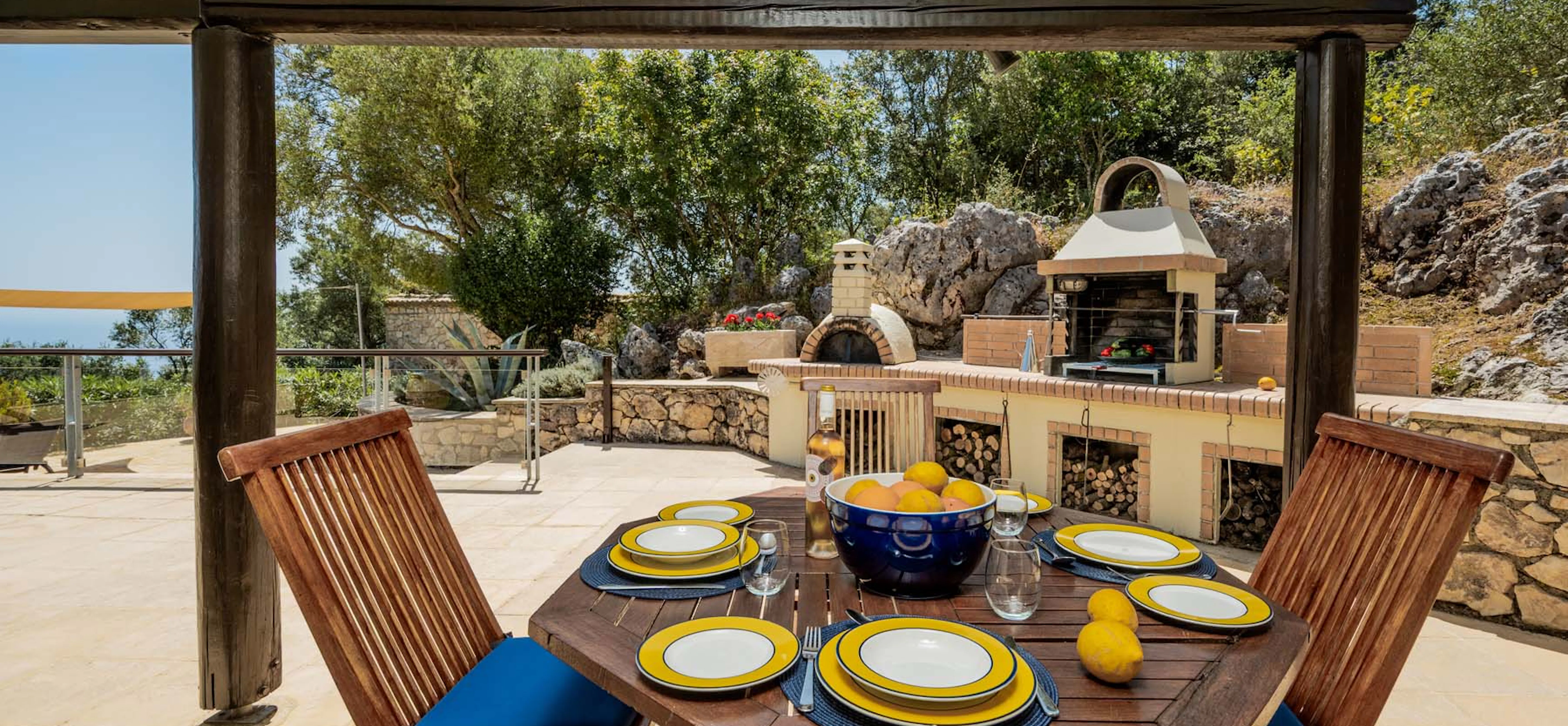 An alfresco dining table set for four with yellow-rimmed crockery, a bowl of citrus fruit, and a wine bottle, beneath a pergola, with the outdoor barbecue station and pizza oven visible in the background.