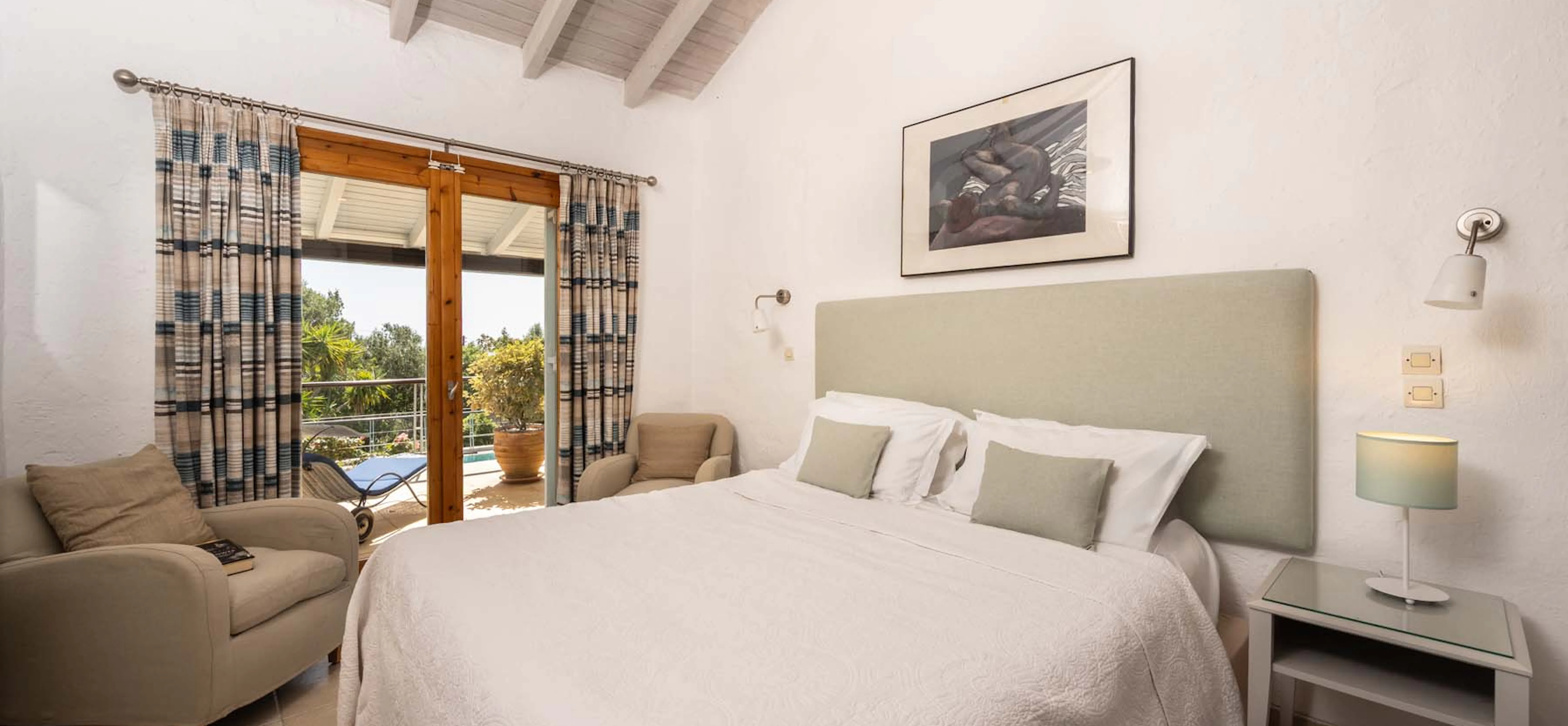 A bright, airy double bedroom with vaulted whitewashed timber ceiling, sage green upholstered headboard, neutral soft furnishings, and French doors opening onto a sunny poolside terrace.
