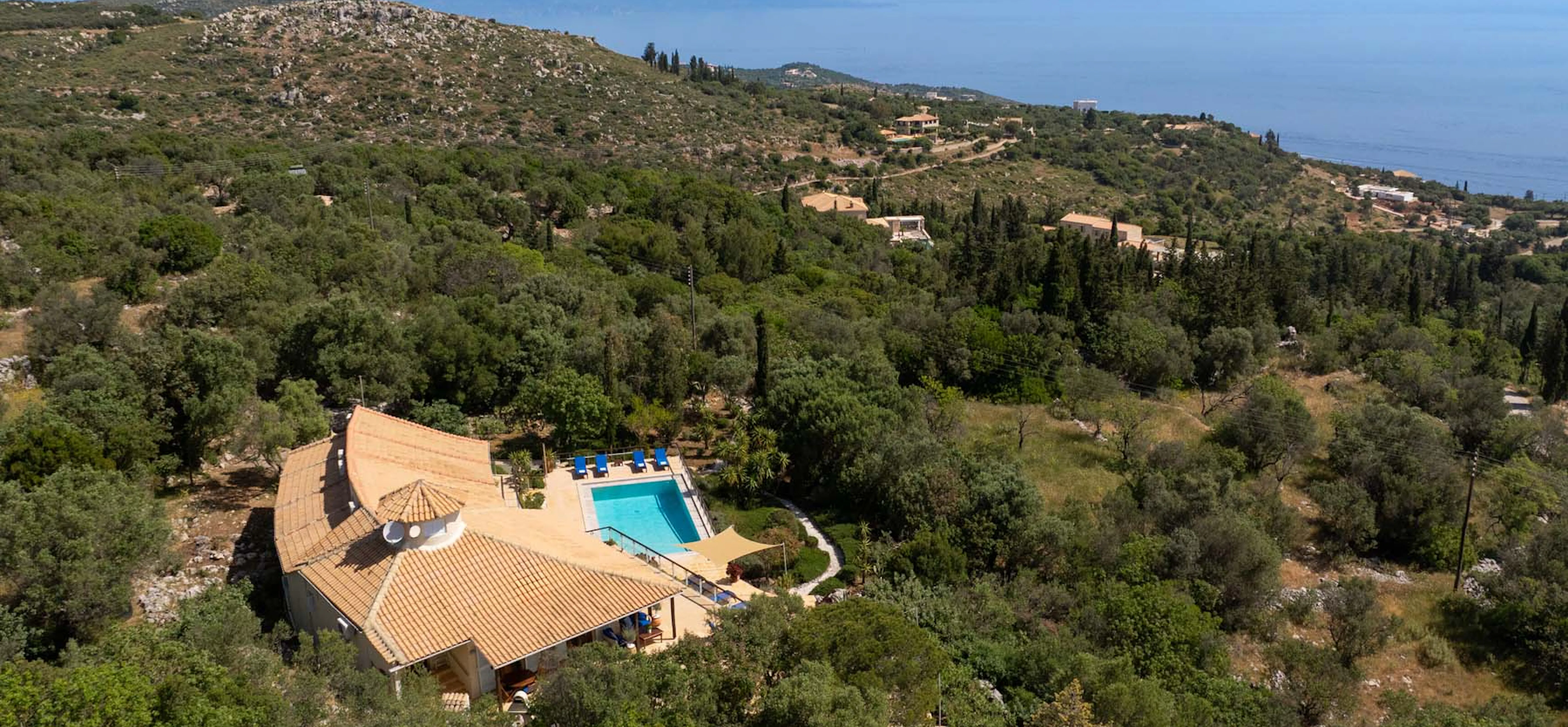 High-altitude aerial view of the villa nestled within an extensive olive grove and Mediterranean scrubland, with the Ionian Sea, distant coastline and mountain ranges visible on the horizon.