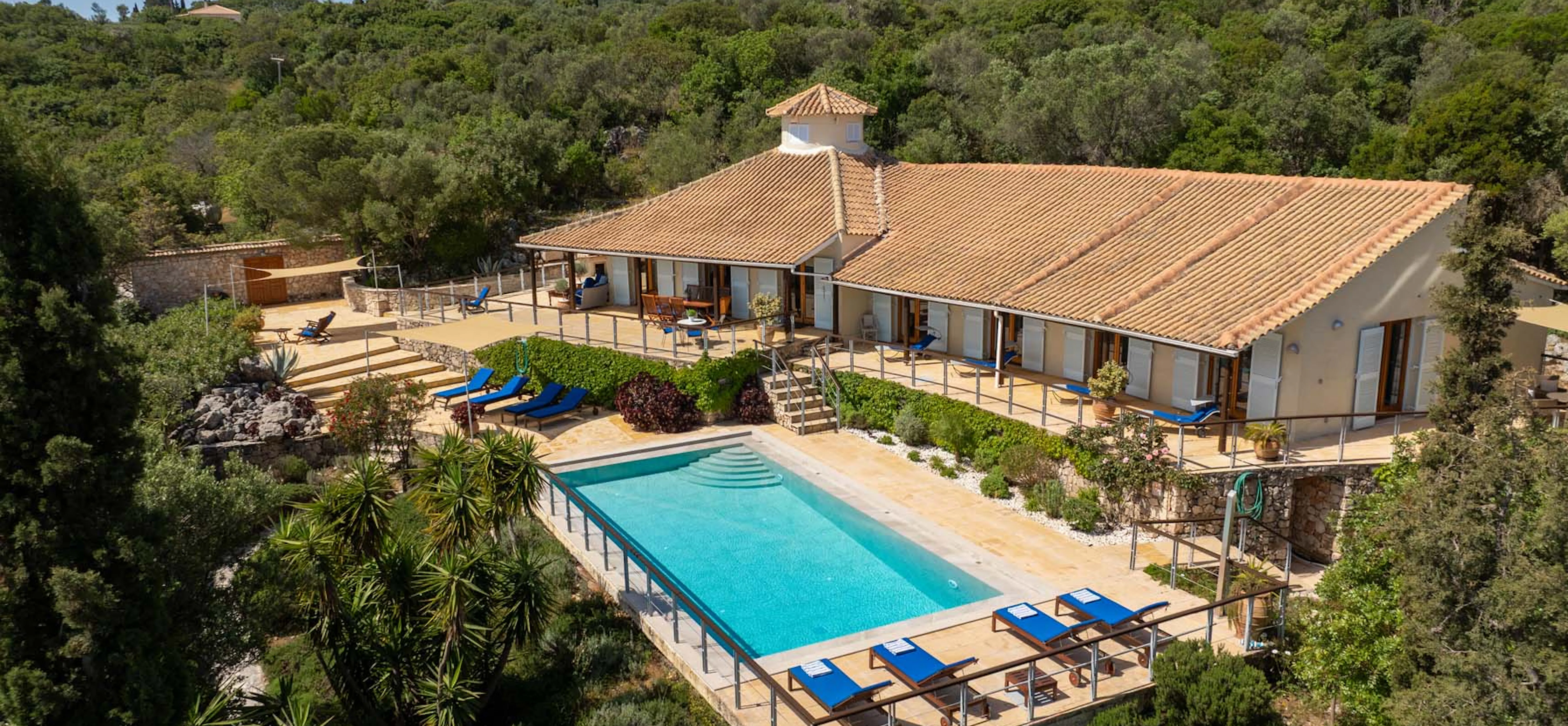 Aerial daytime view of the villa from a higher angle, showing the full pool terrace with blue sun loungers, landscaped gardens, multiple outdoor seating areas, and the surrounding wooded hillside.