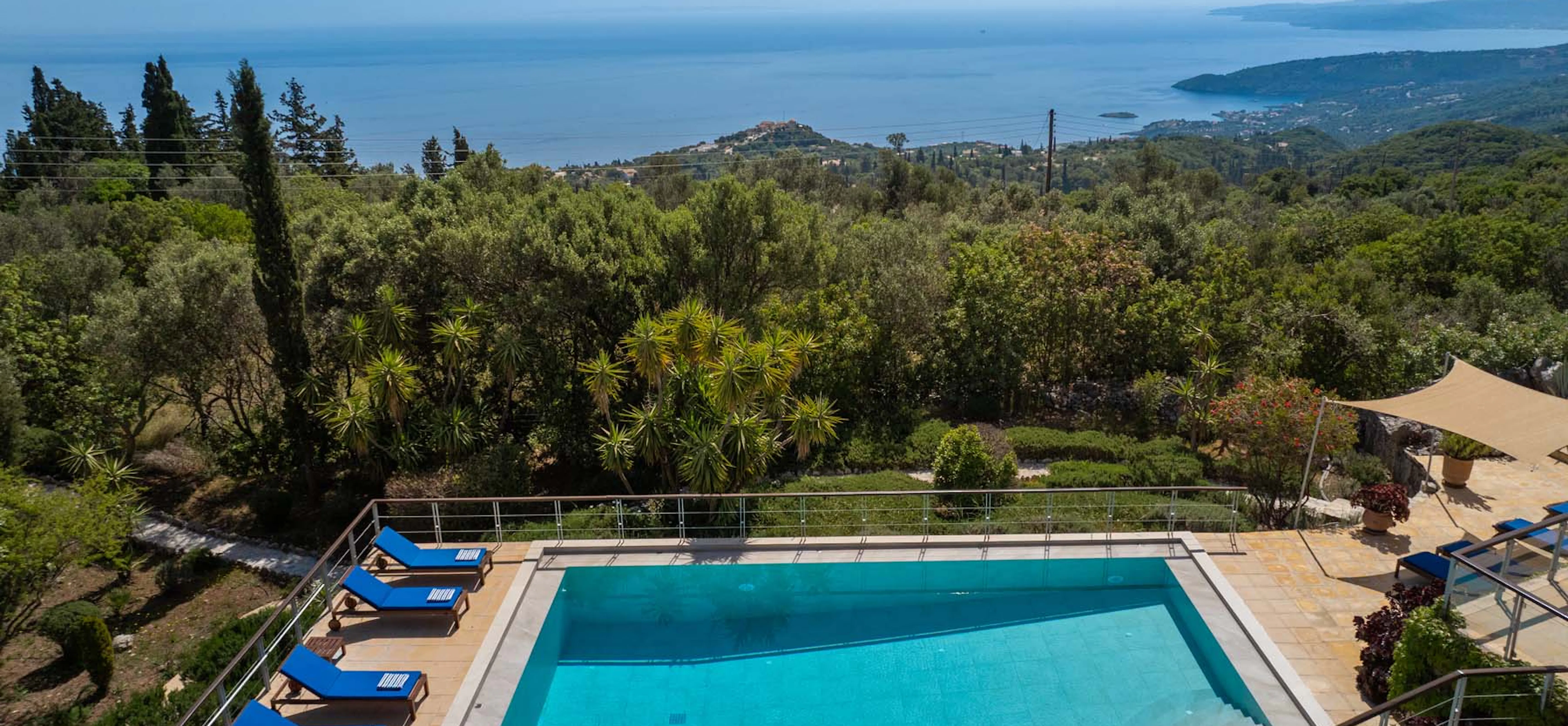 Elevated drone view looking over the villa's rectangular swimming pool with blue sun loungers, across landscaped gardens of yucca and flowering shrubs, to a spectacular wide panorama of the Ionian Sea, a coastal headland, and distant Greek islands.